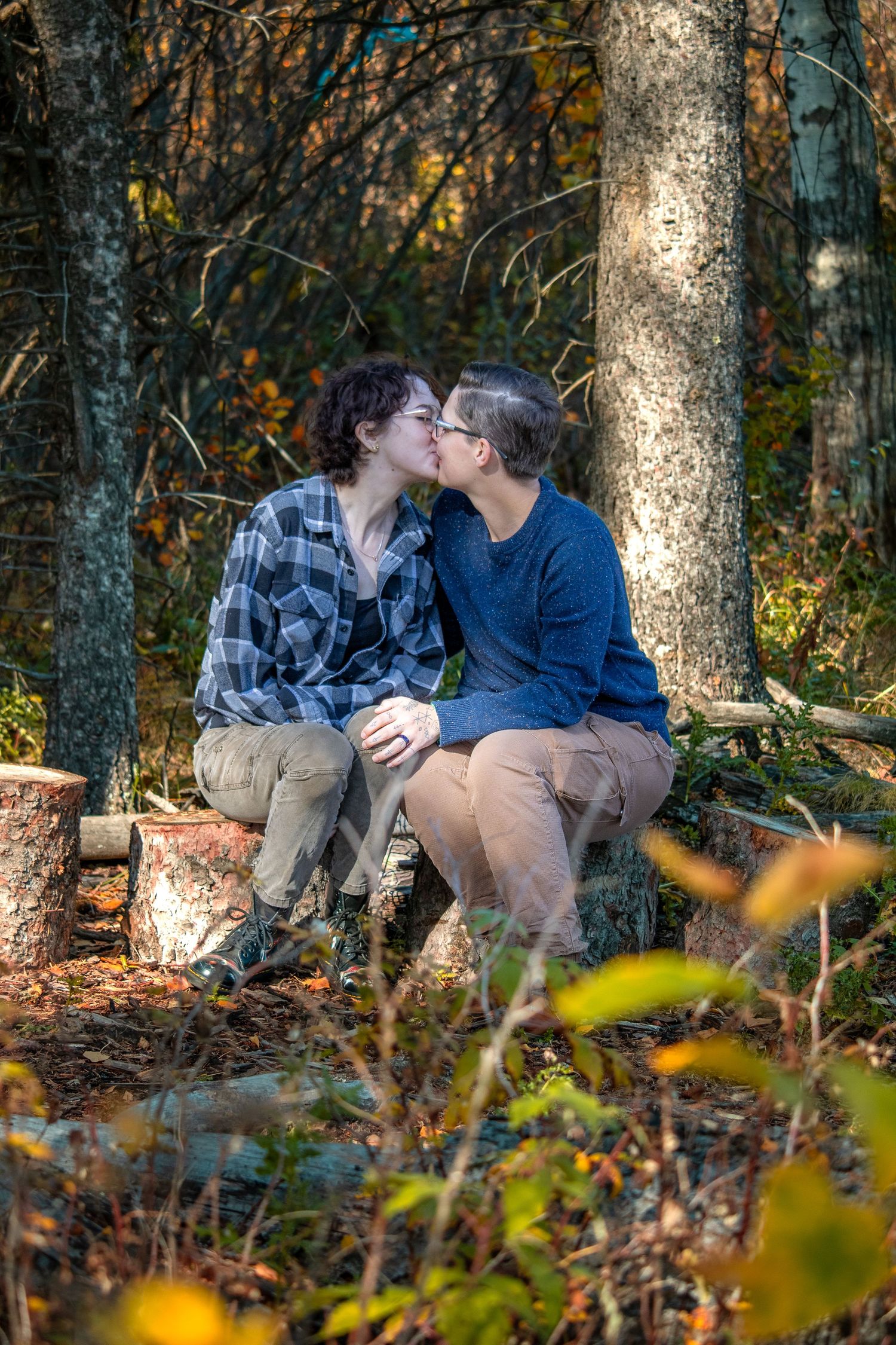A romantic couple shares a tender kiss while sitting on a log in an autumn forest setting.