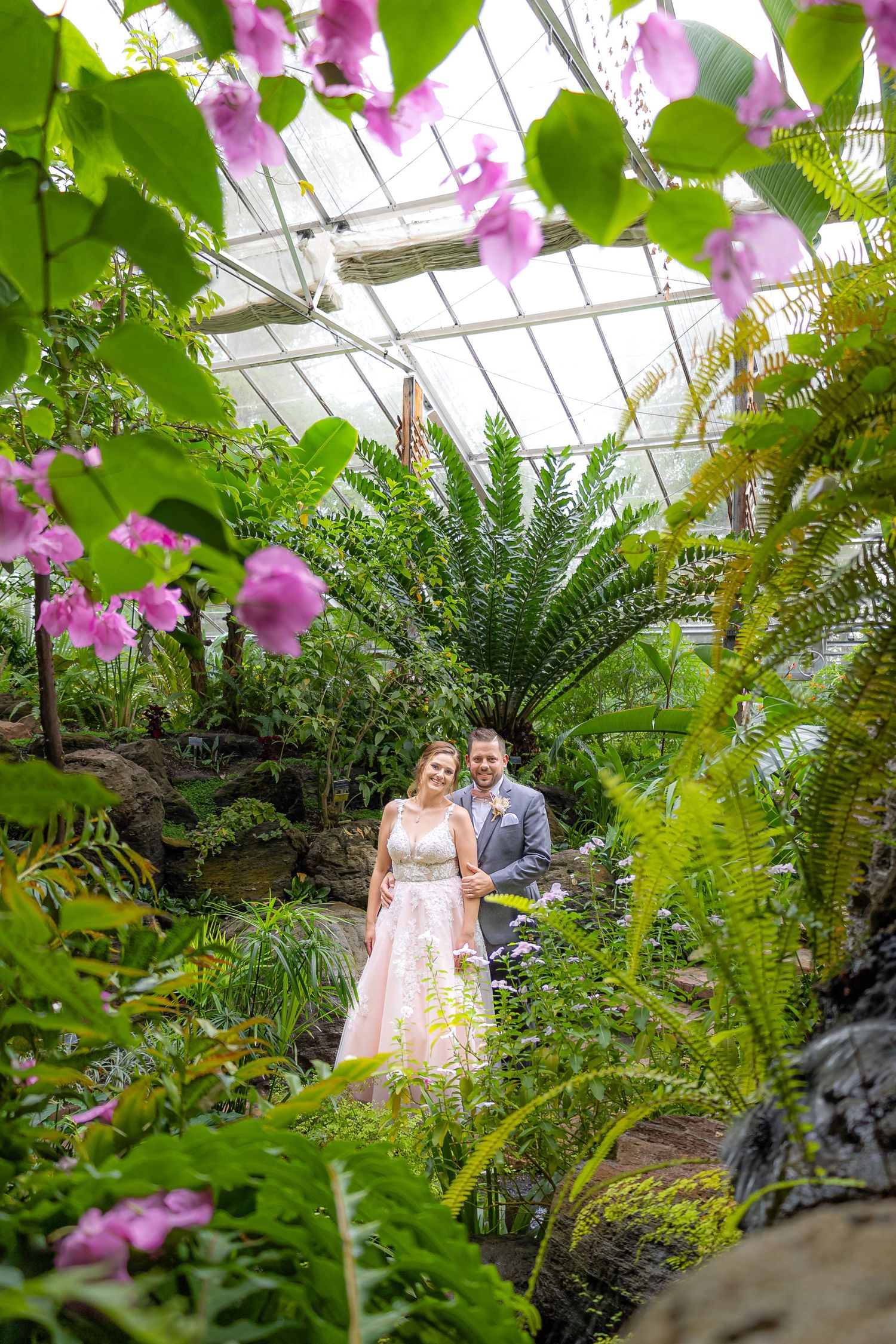 A couple poses in a lush greenhouse setting with pink flowers and tropical plants.