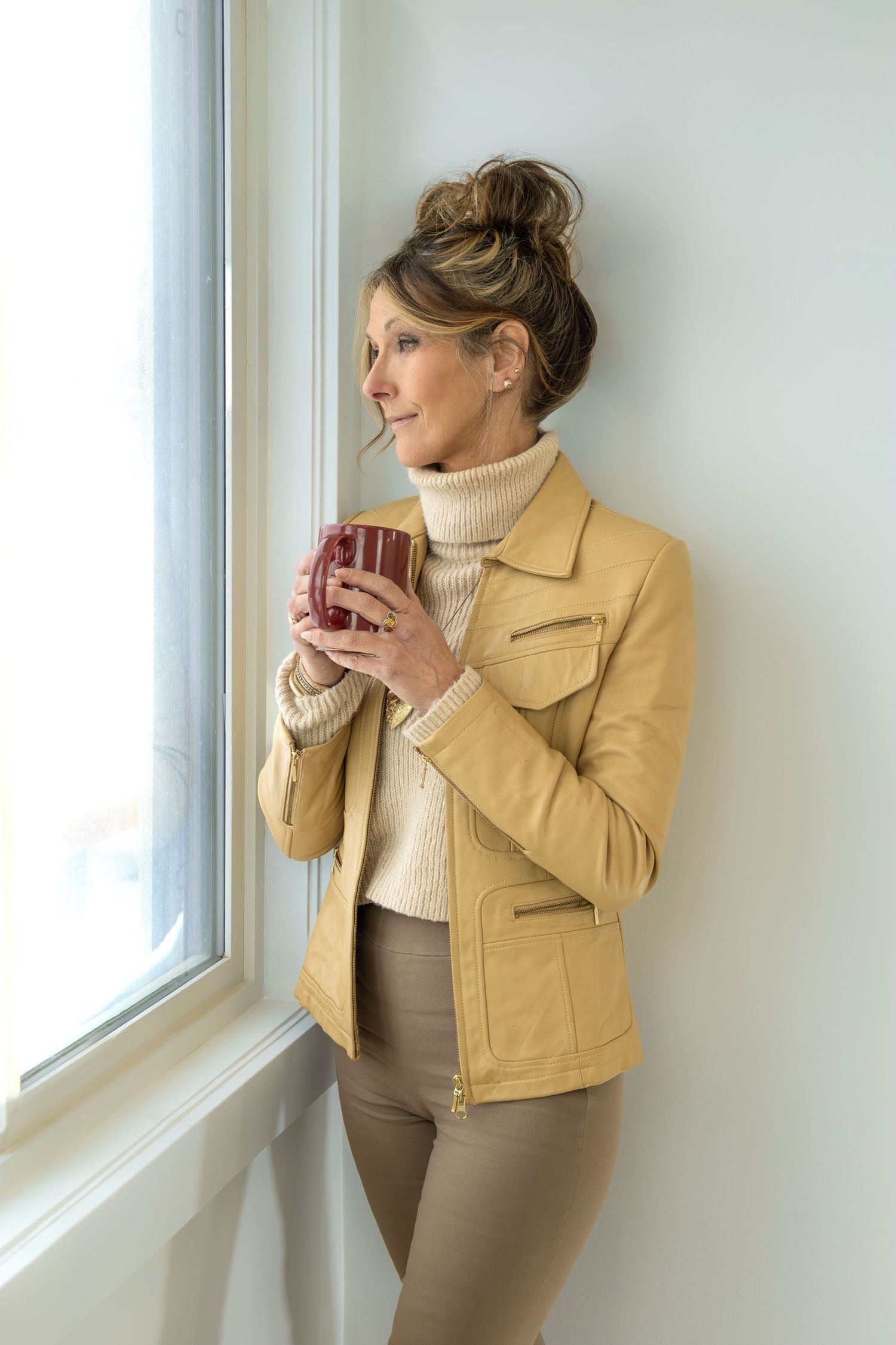 A woman in a tan jacket and cream turtleneck gazes out a window while holding a mug dreaming of the warmer days