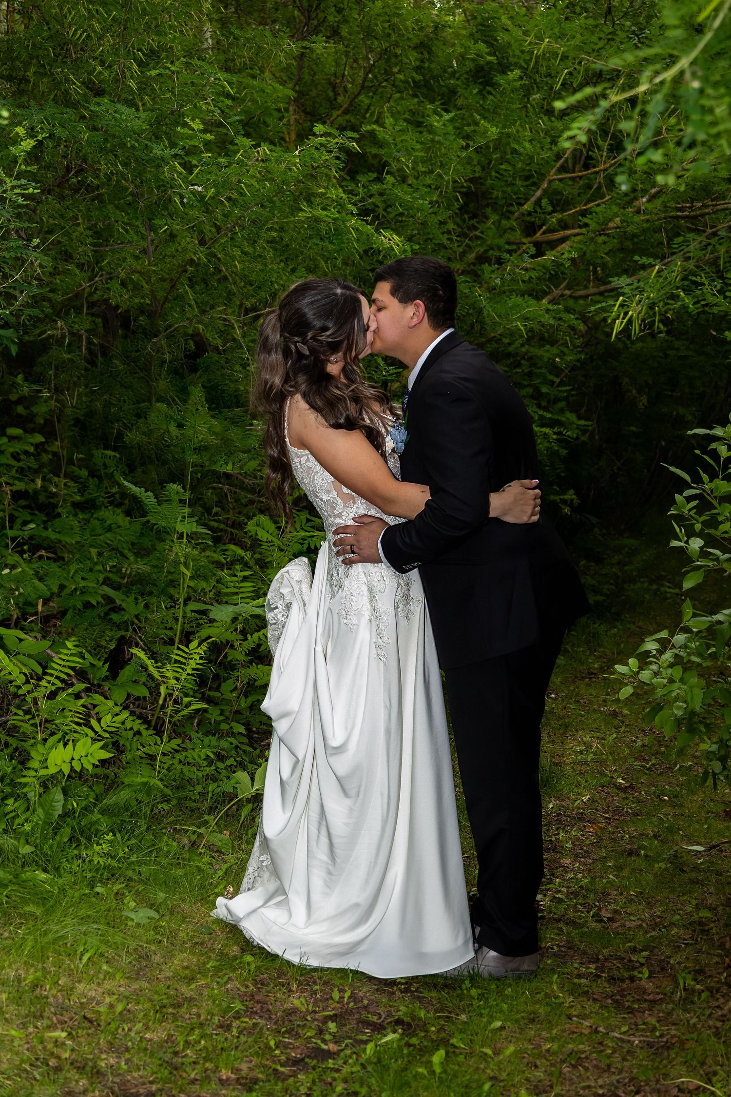 A couple shares an intimate moment in formal attire among lush green foliage during an outdoor wedding photoshoot.