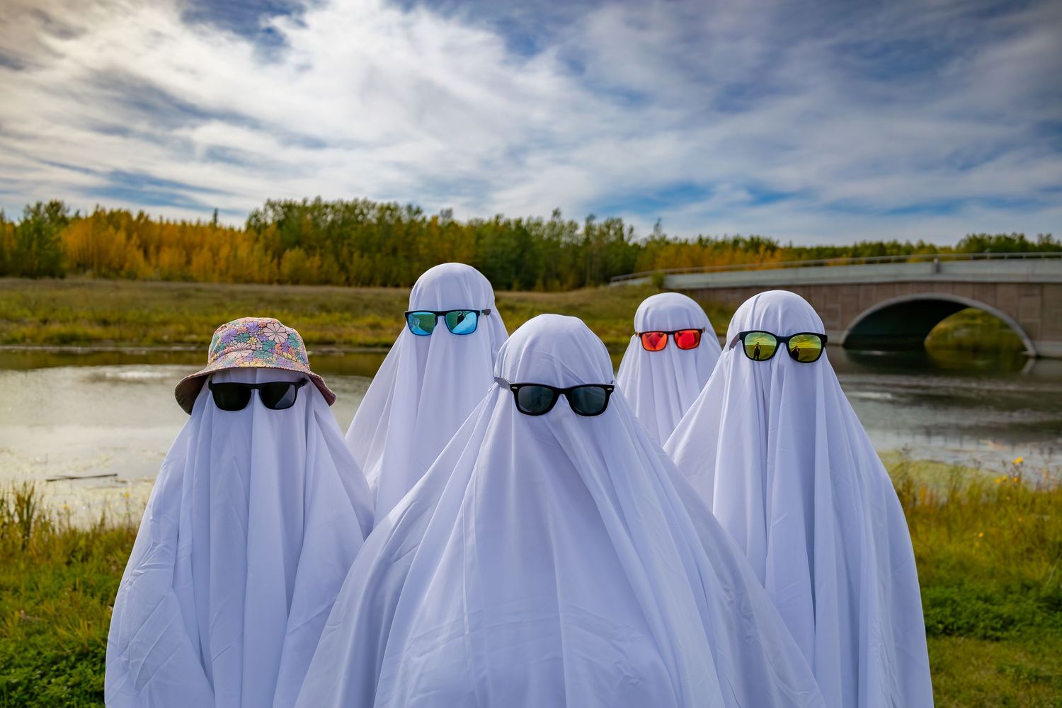 Five figures dressed as ghosts wearing sunglasses pose by a river under cloudy skies.