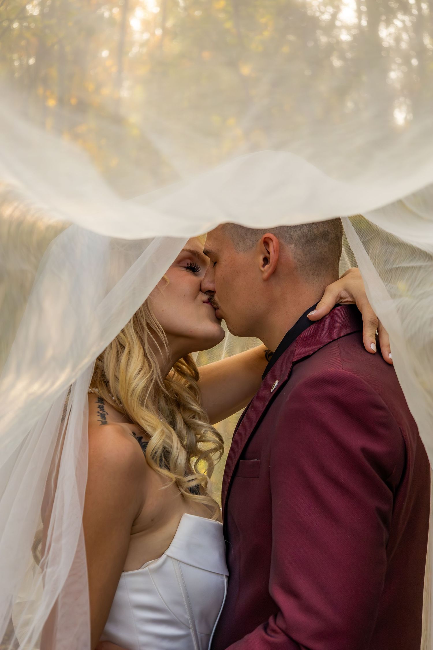A romantic wedding couple shares an intimate kiss under a flowing white veil against a soft, dreamy background.