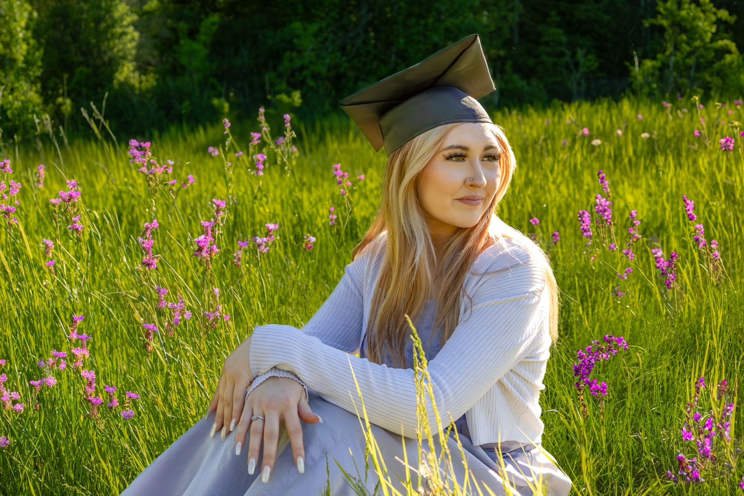 Graduate in cap and gown sits thoughtfully in a summer meadow with pink wildflowers blooming.