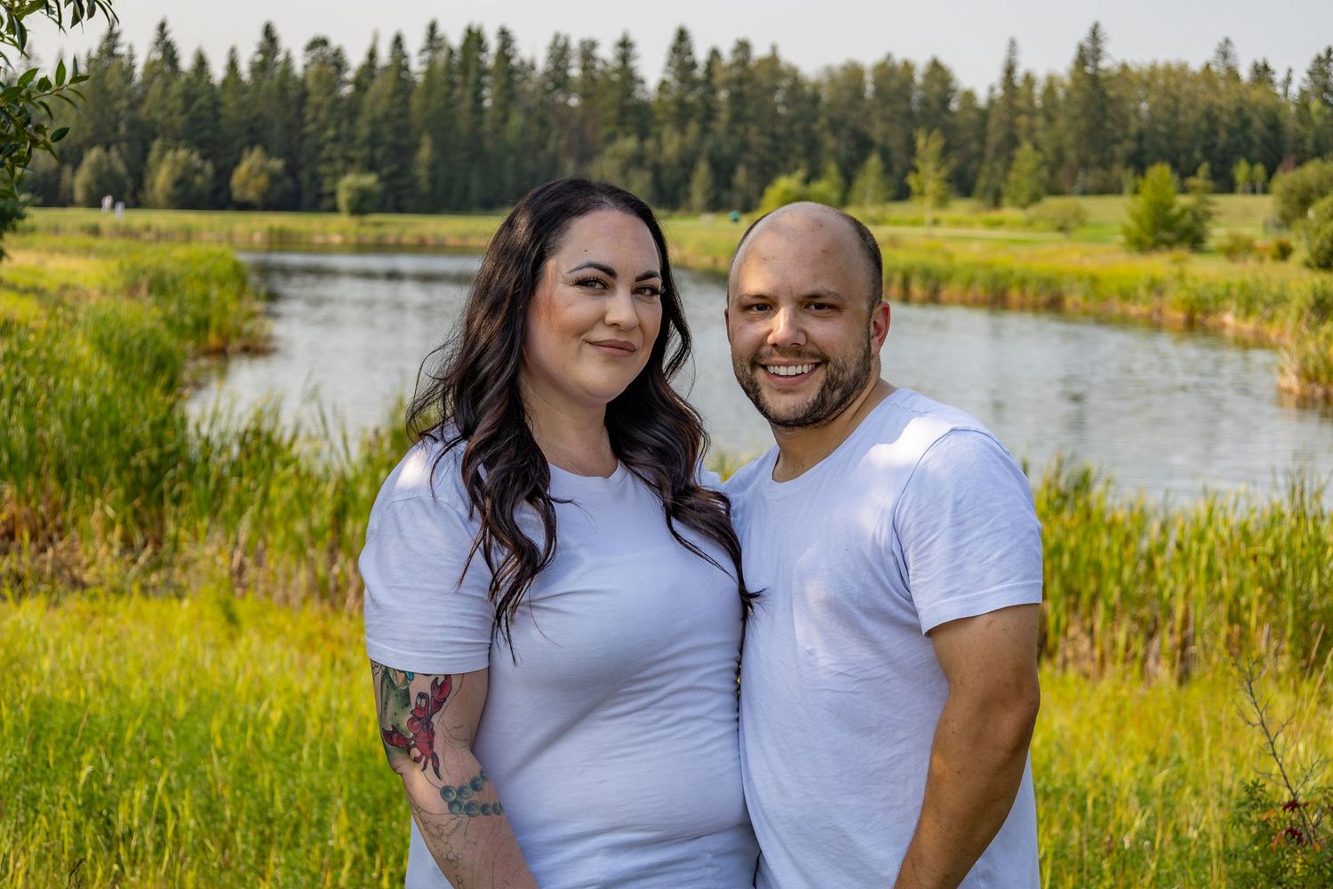 A couple poses together by a scenic lake surrounded by evergreen trees and tall grass.