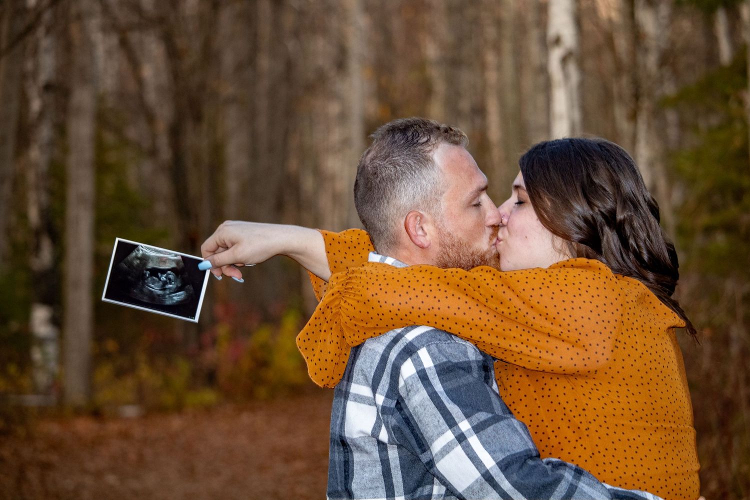 Couple sharing intimate moment while holding ultrasound photo in forest setting.
