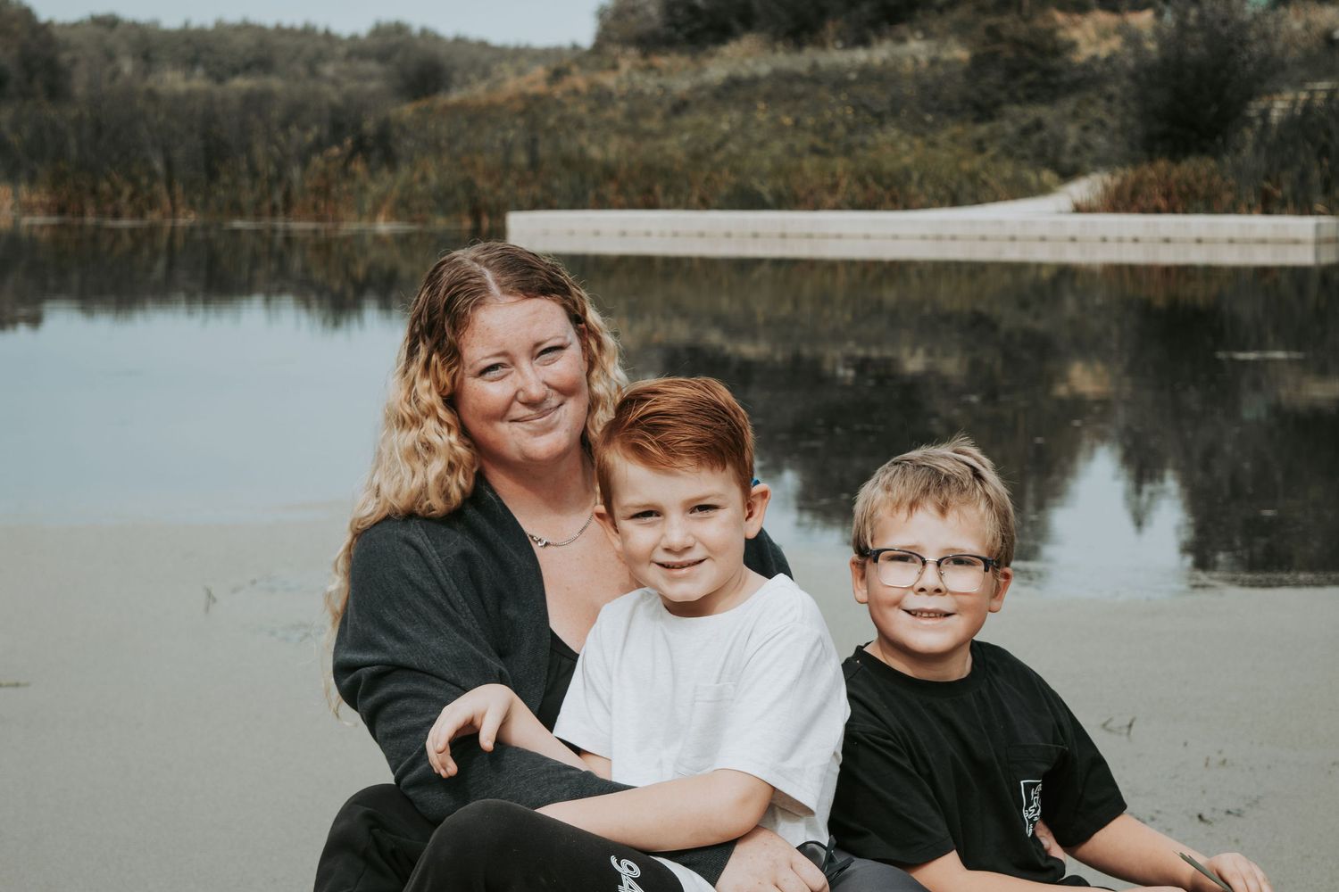 A heartwarming lakeside portrait of a parent with twin boys sitting together at the water's edge.