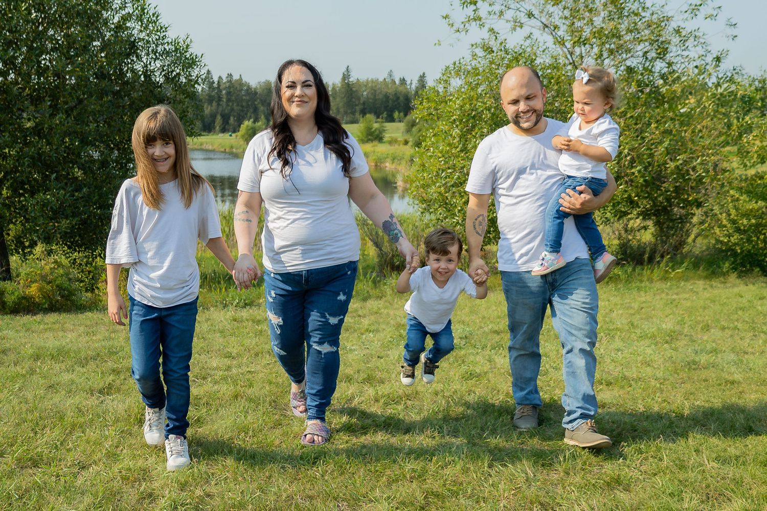 Family walks together on grassy path near lake during summer photo session.