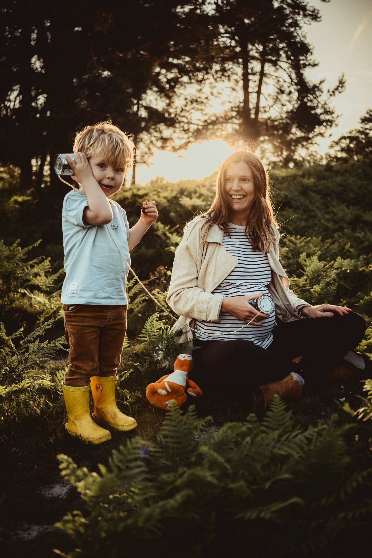 playful moment between a toddler and his pregnant mum using a tin can phone at sunset in the countryside