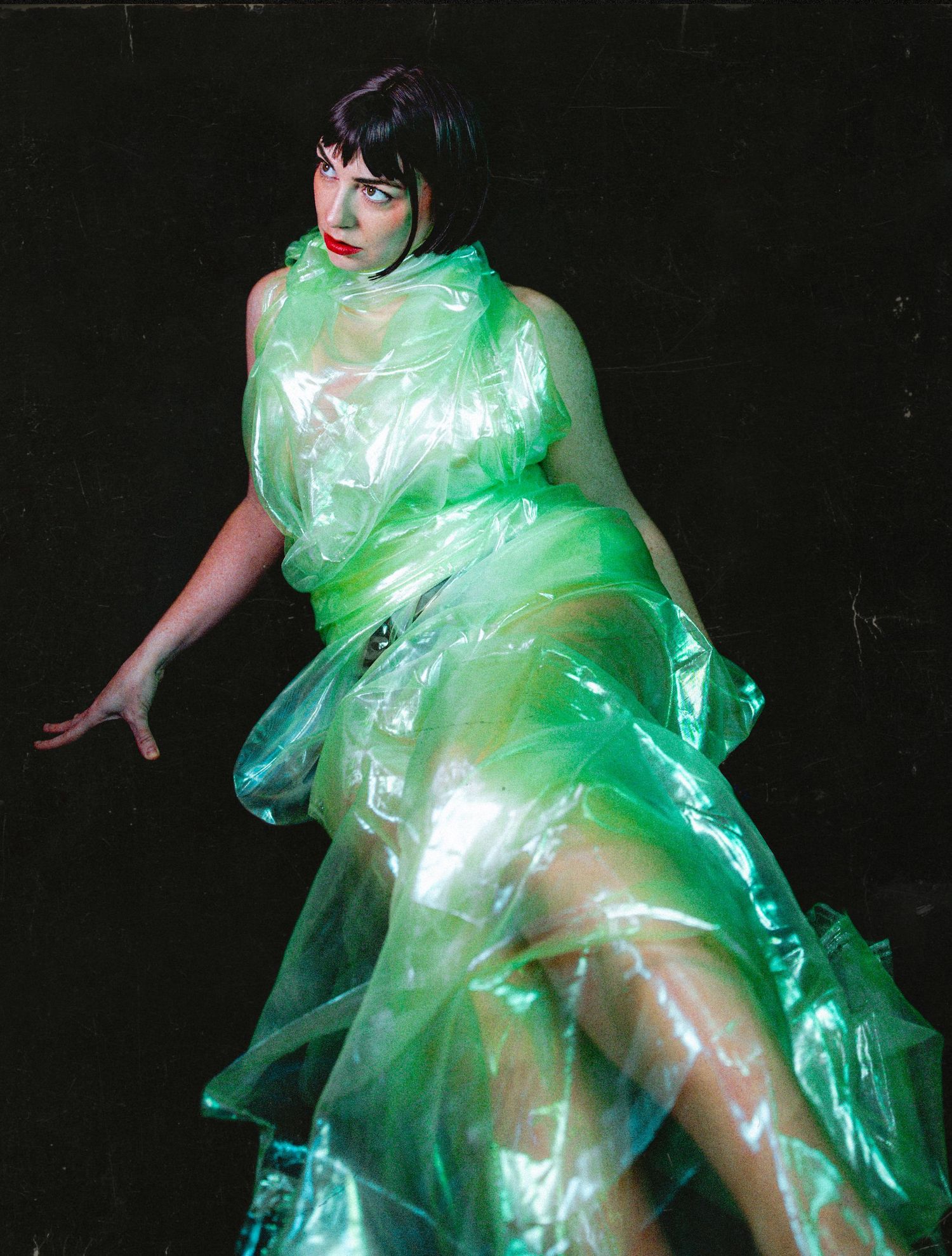 Figure in translucent green plastic dress poses dramatically against dark background with ethereal lighting effects.