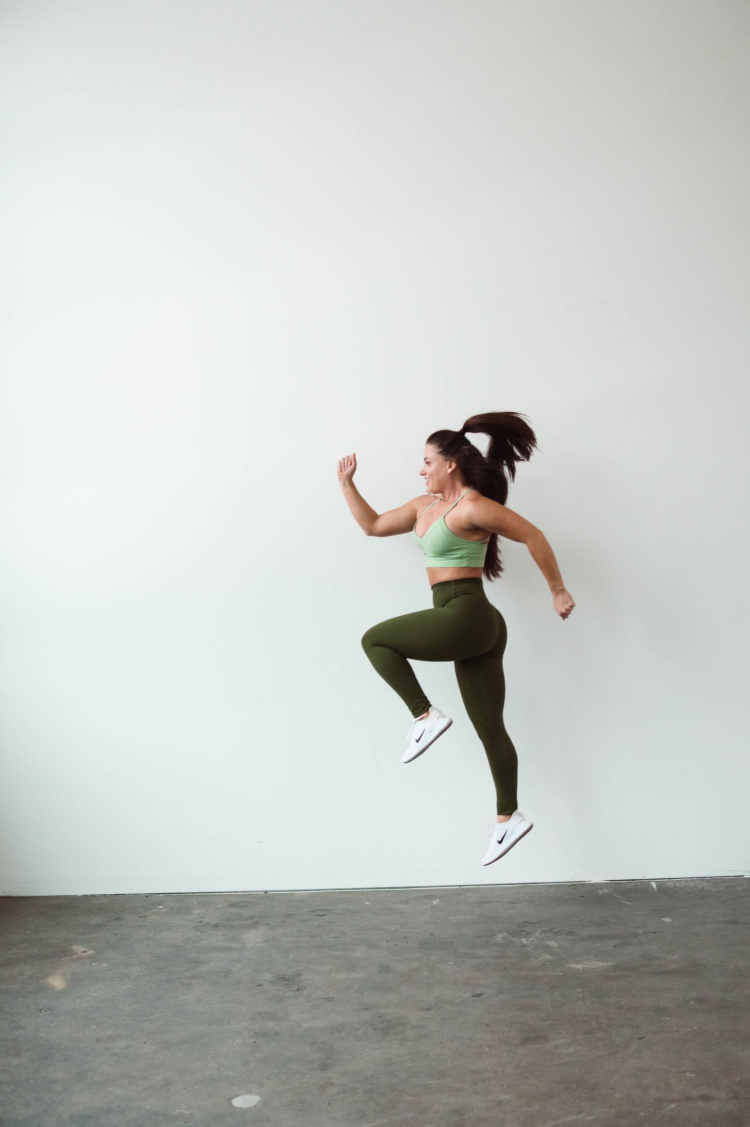 Athletic figure in olive green workout attire jumping energetically against white wall.