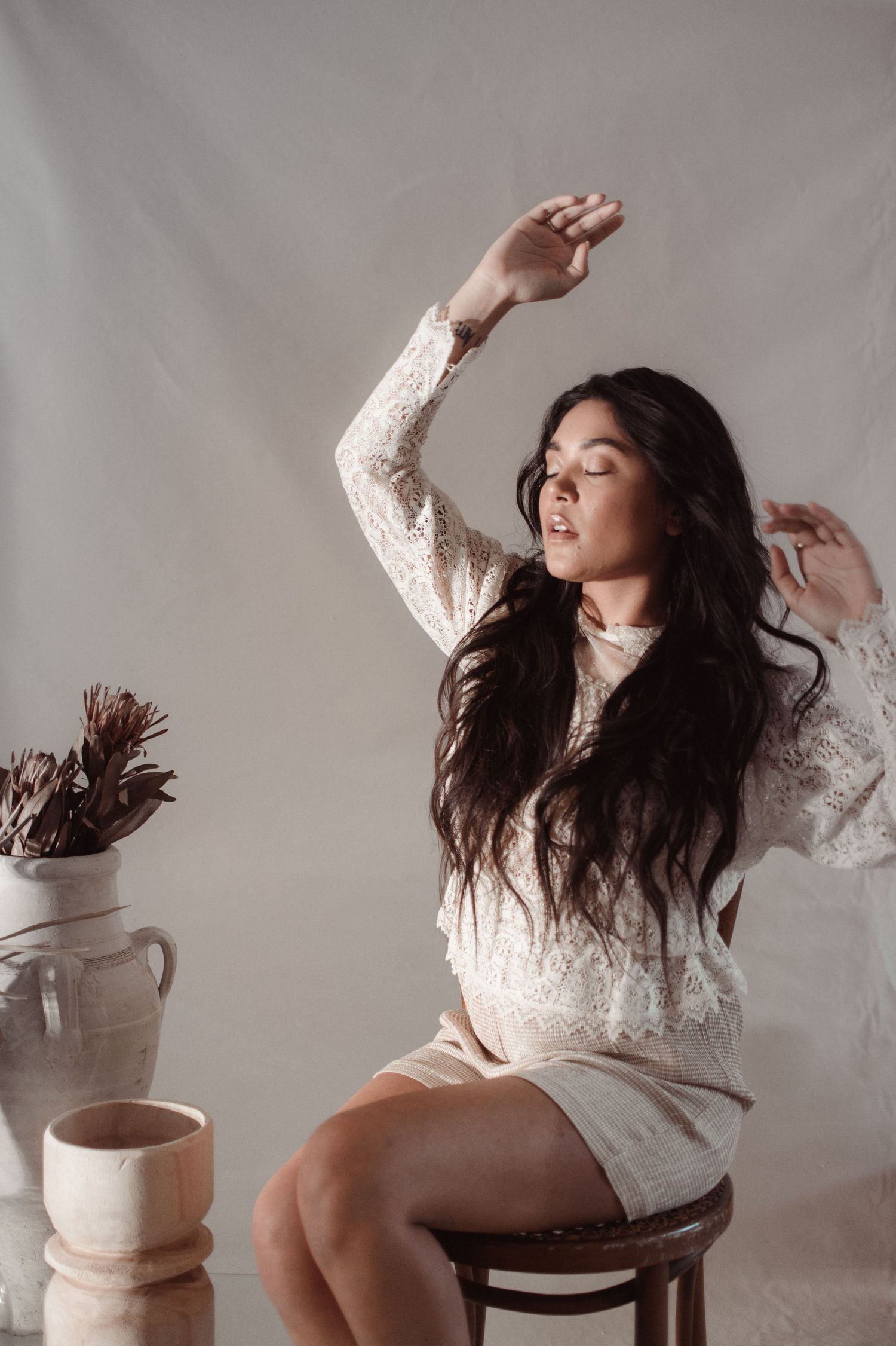 Woman in white lace dress poses gracefully on wooden chair with coffee cup nearby.