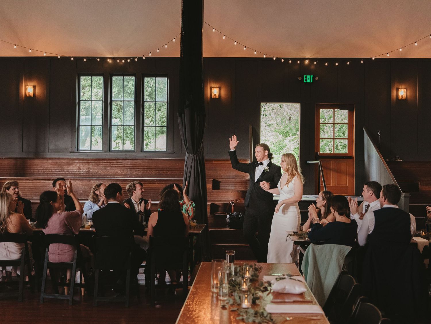 Wedding reception in a rustic wooden venue with string lights and guests seated at long tables.