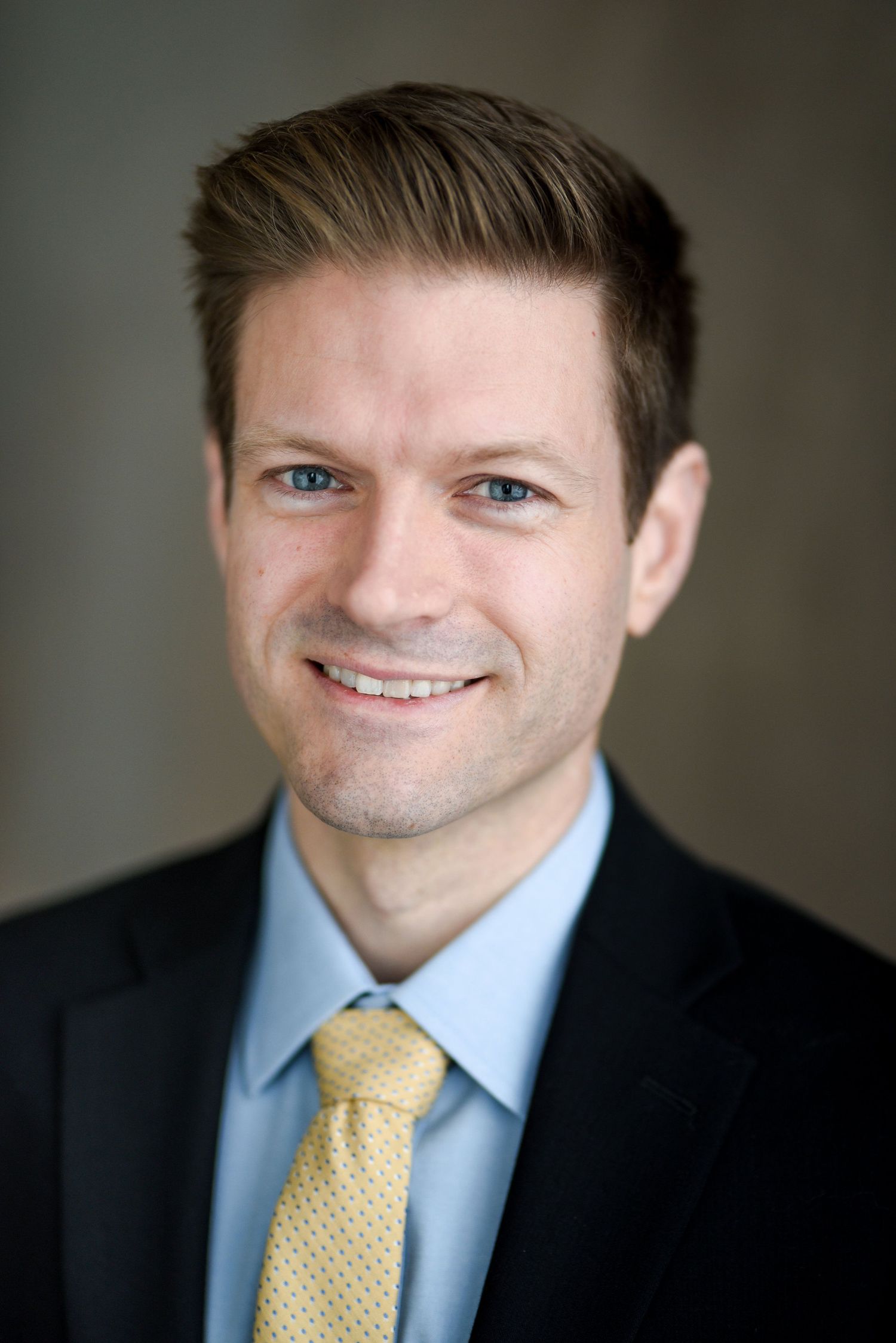 Professional headshot showing person in dark suit with light blue dress shirt and yellow tie against blurred background.