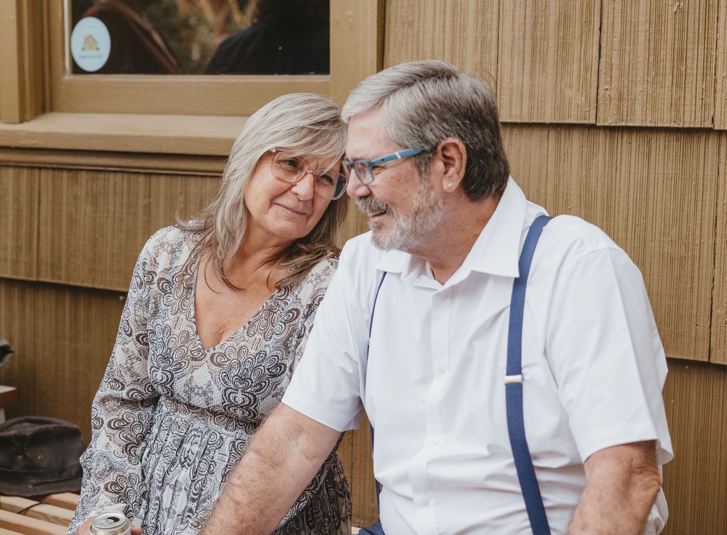 An elderly couple sits together on outdoor wooden furniture while looking at a mobile device.