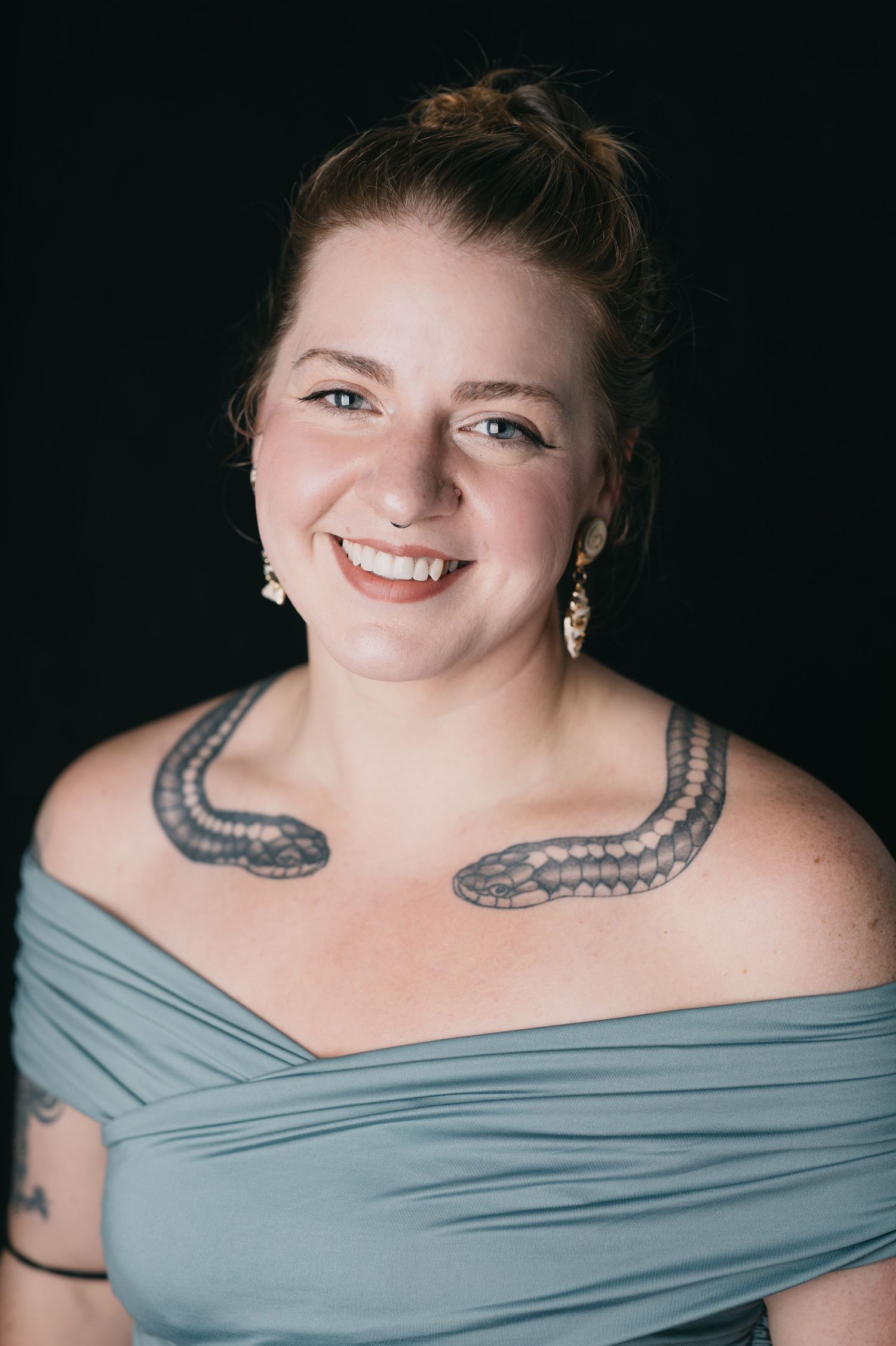Portrait of a person in an off-shoulder grey dress with visible shoulder tattoos.