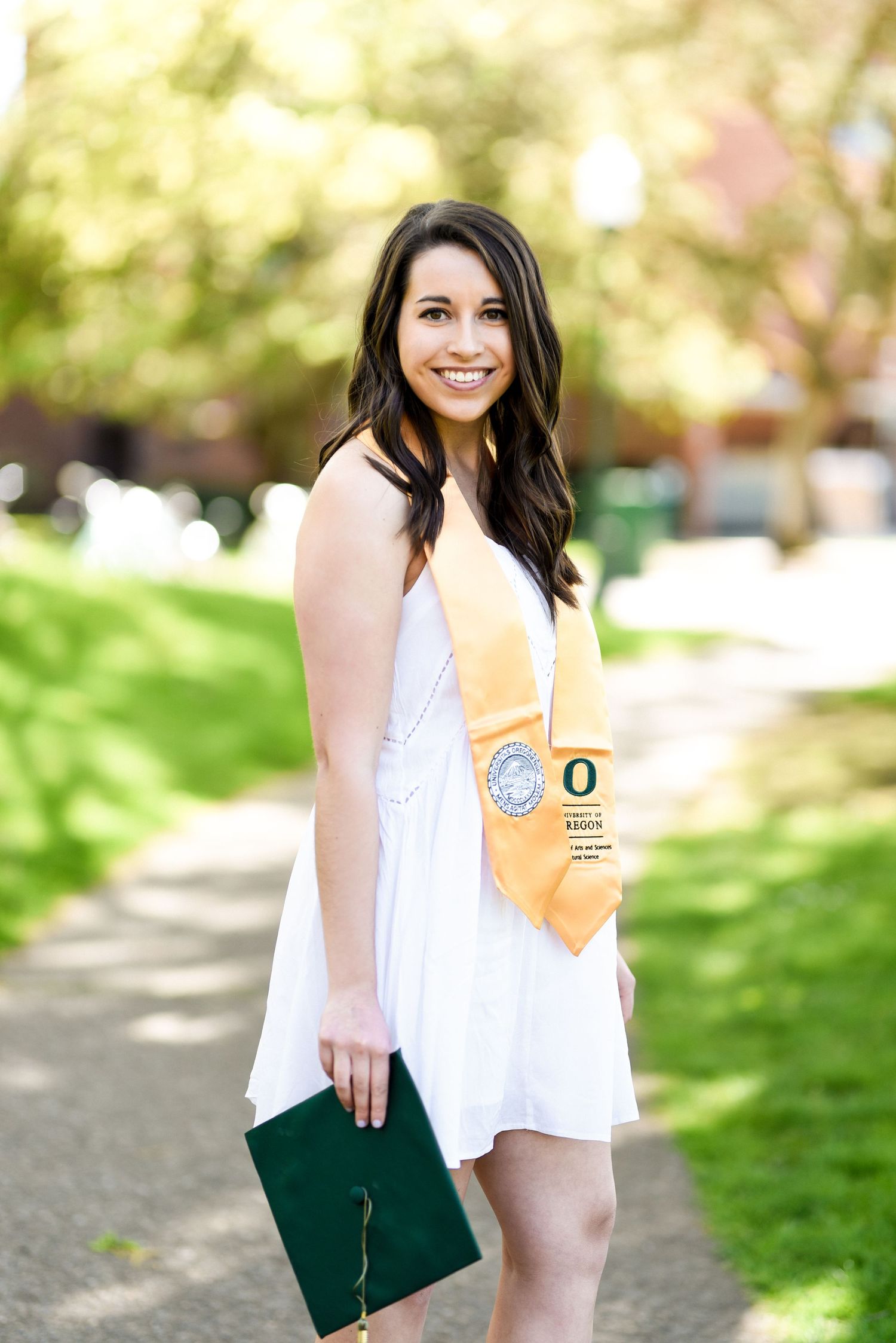 University of Oregon graduate in white dress with yellow sash holding diploma.