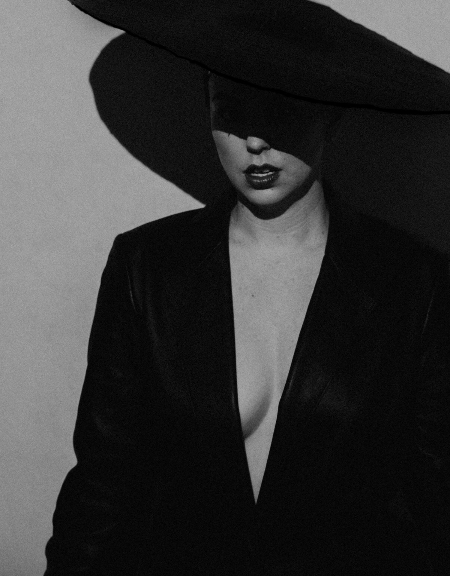 Dramatic black and white portrait featuring a wide-brimmed hat, red lips, and plunging black blazer against light background.