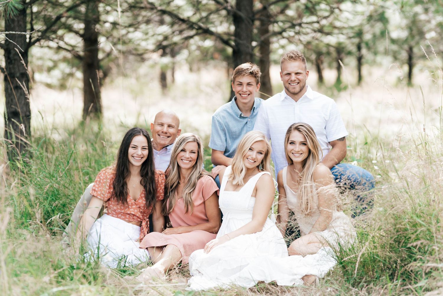 A group poses together in a grassy field surrounded by pine trees during a casual outdoor photoshoot.