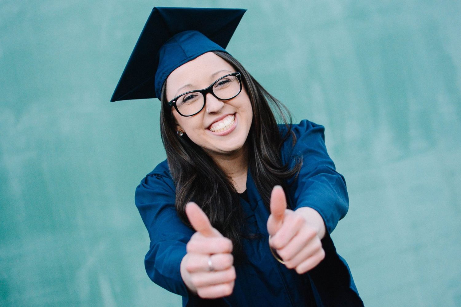 Graduate in blue cap and gown gives enthusiastic thumbs up against teal background.