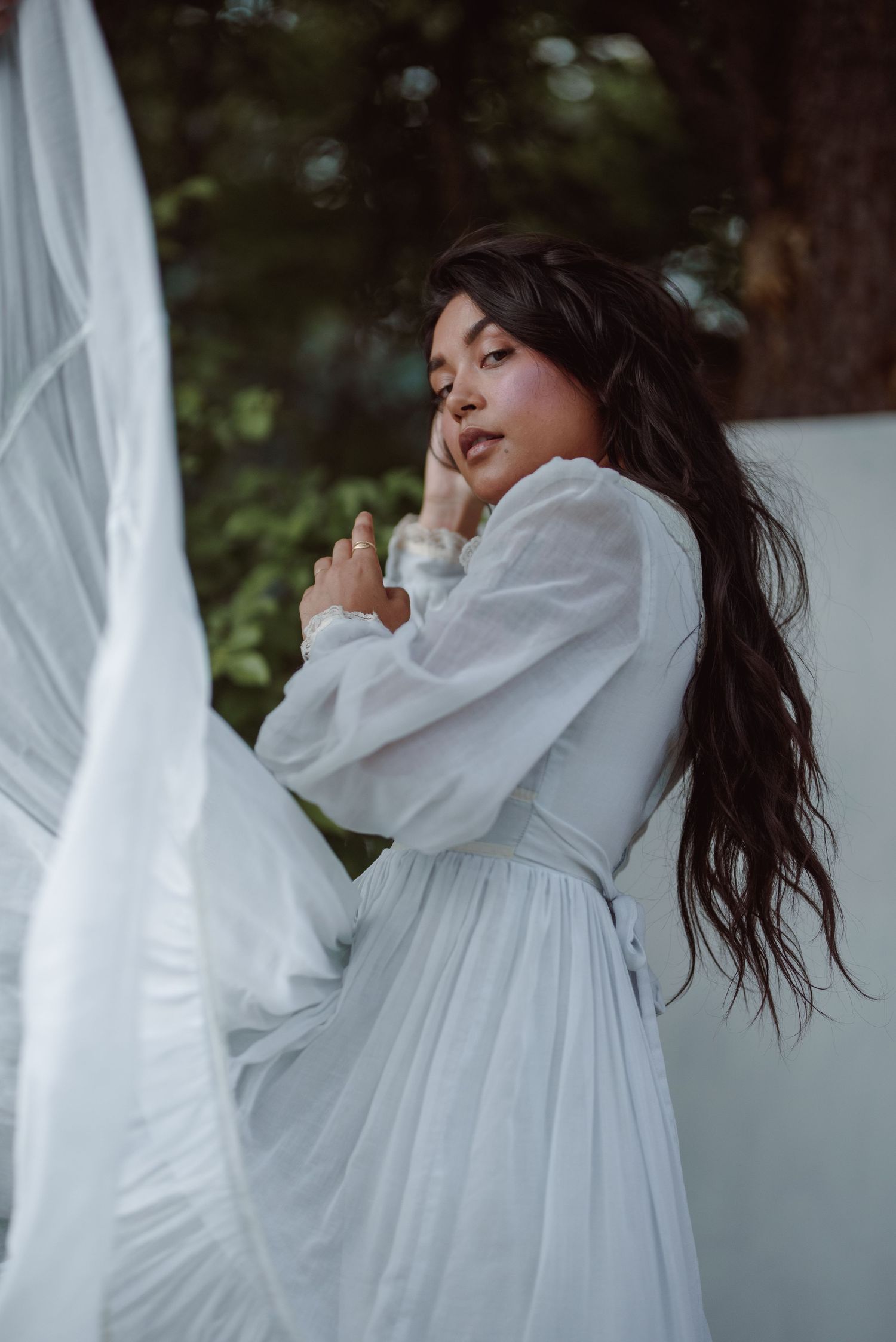 Woman in flowing white dress poses ethereally in garden setting with billowing fabric.