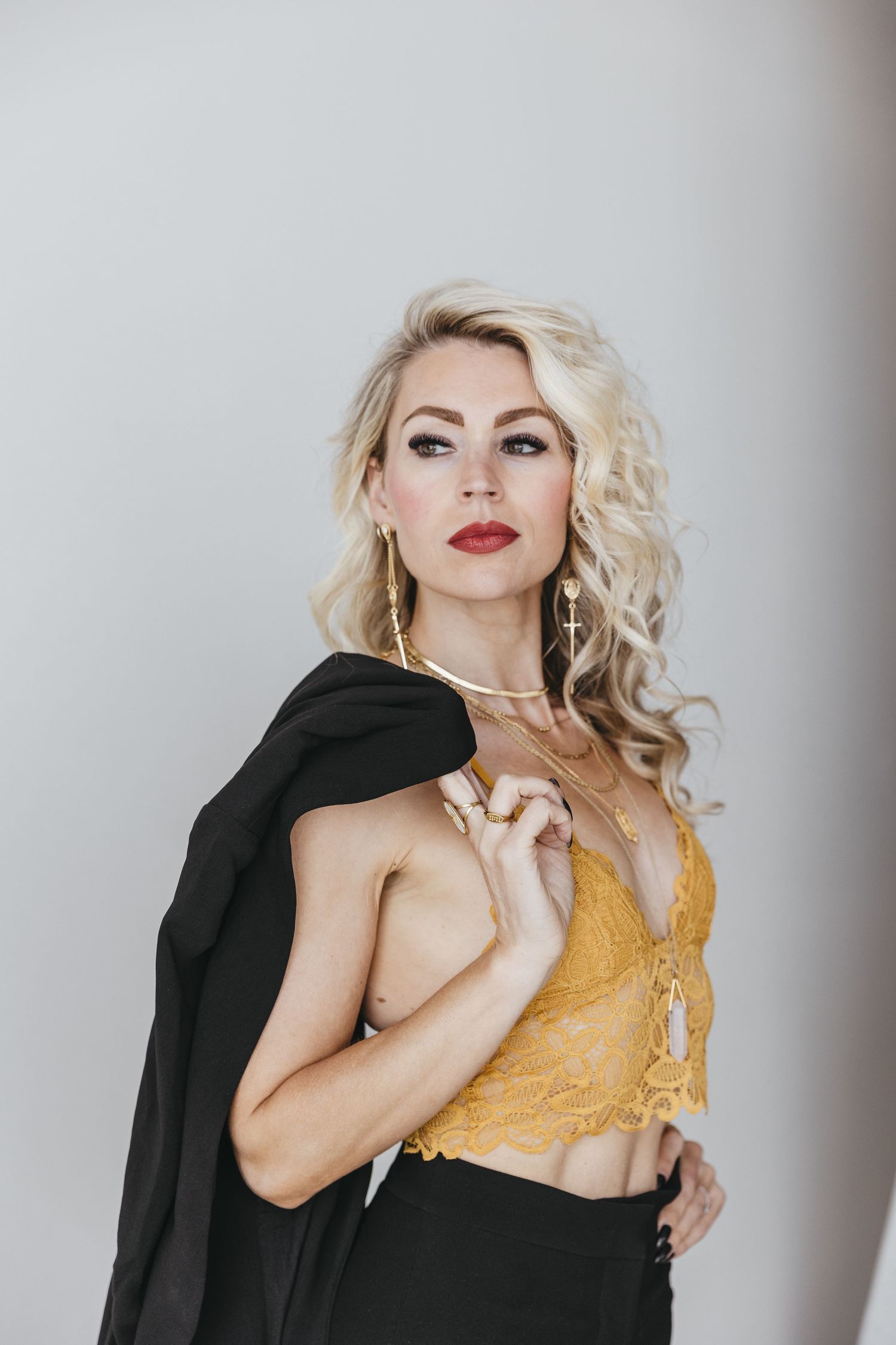 Stylish portrait featuring blonde wavy hair, yellow lace crop top, and black jacket against neutral background.
