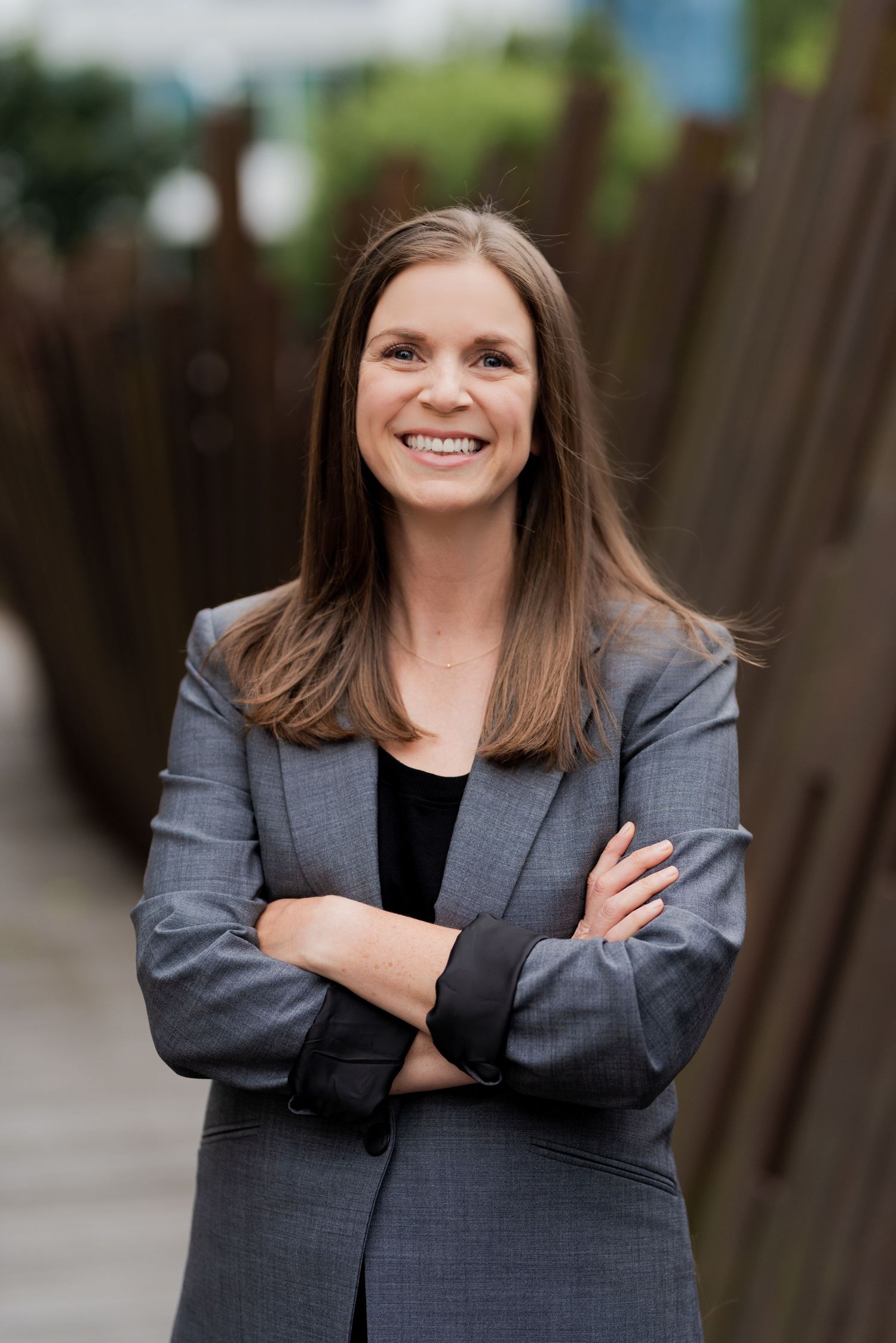 Professional portrait of person in grey suit jacket standing with arms crossed outdoors.