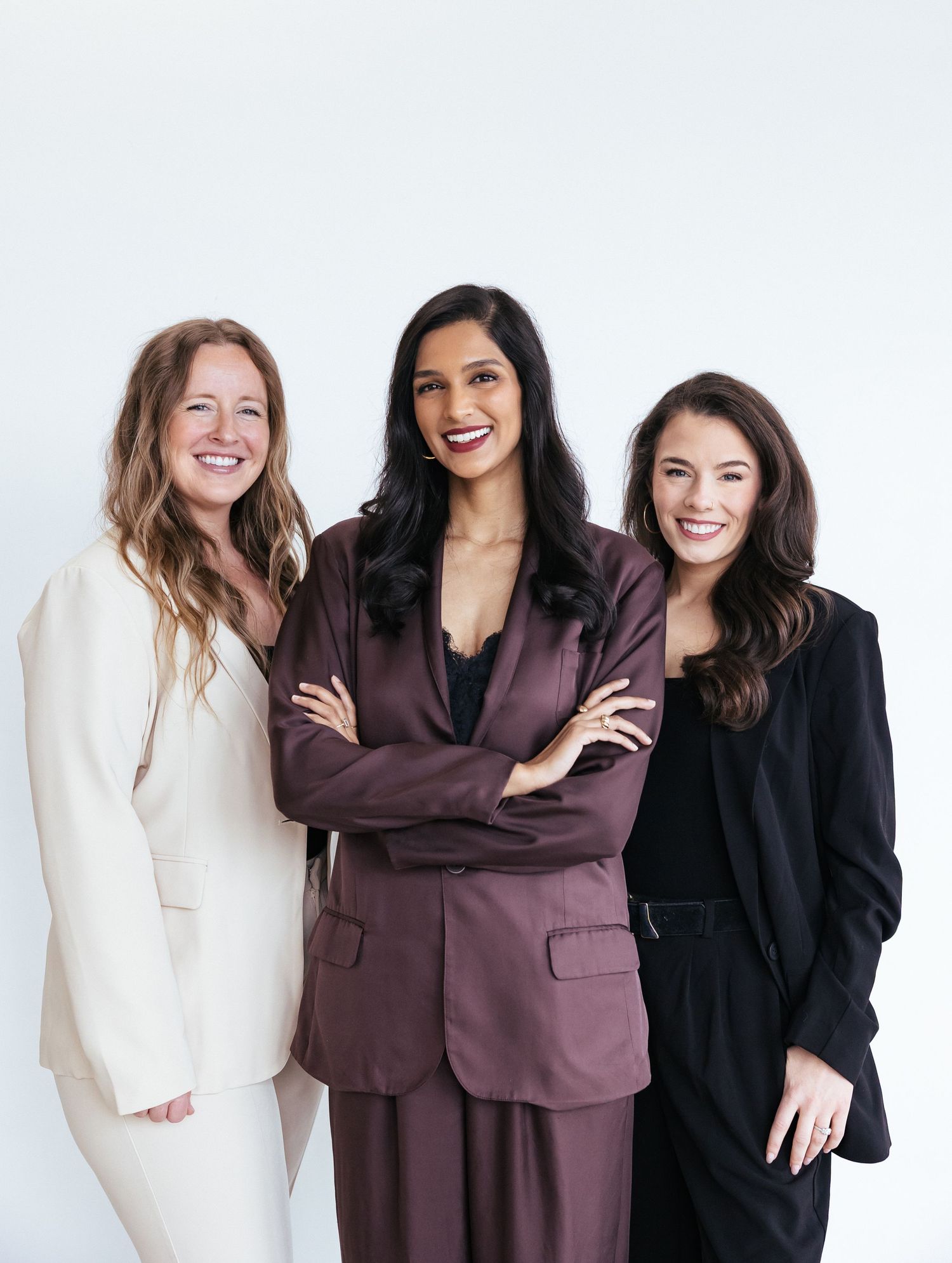 Three professional women in blazers smiling confidently, posing together against a white background.