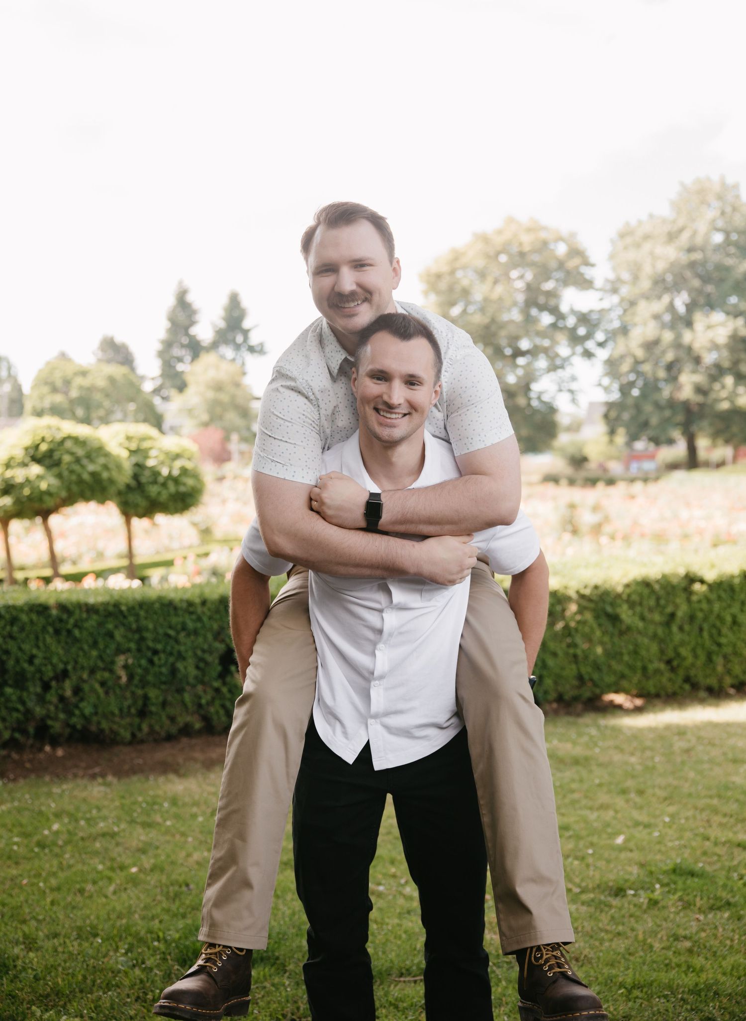 A candid outdoor portrait of two people embracing and smiling in a garden setting with trees and hedges in the background.