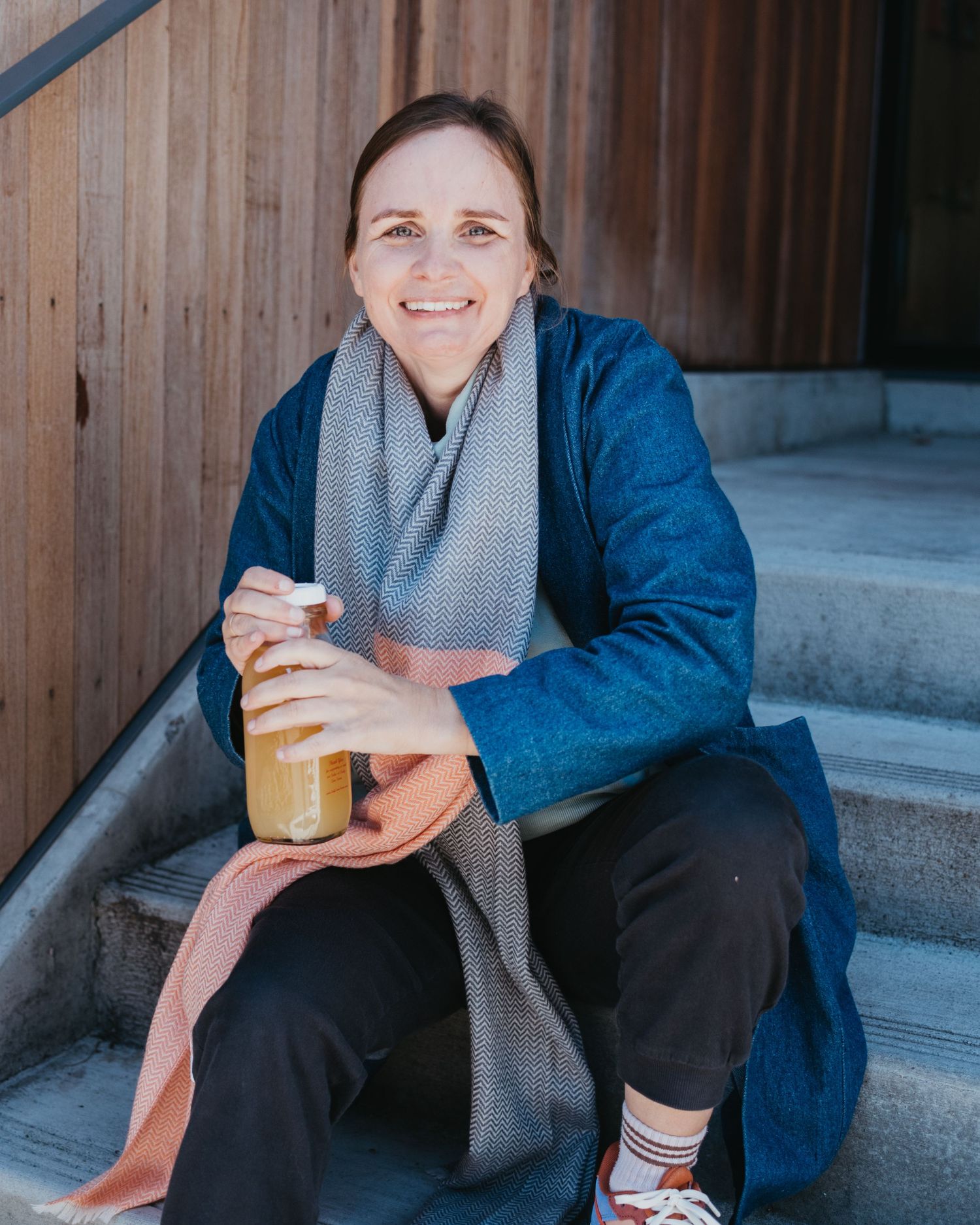 Person in blue coat and scarf sitting on steps with beverage.