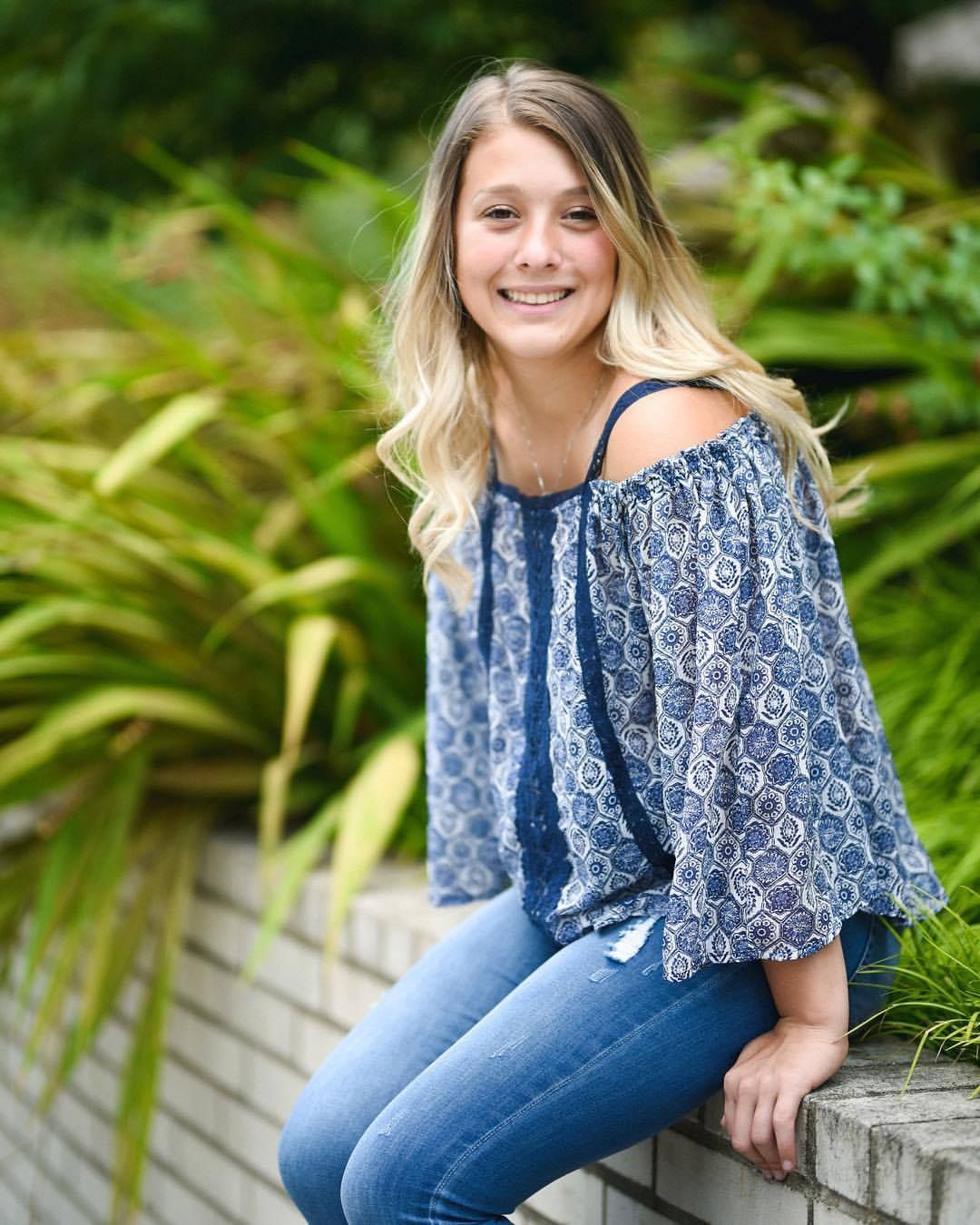 Casual portrait of woman in blue patterned top sitting on stone wall with tropical plants.