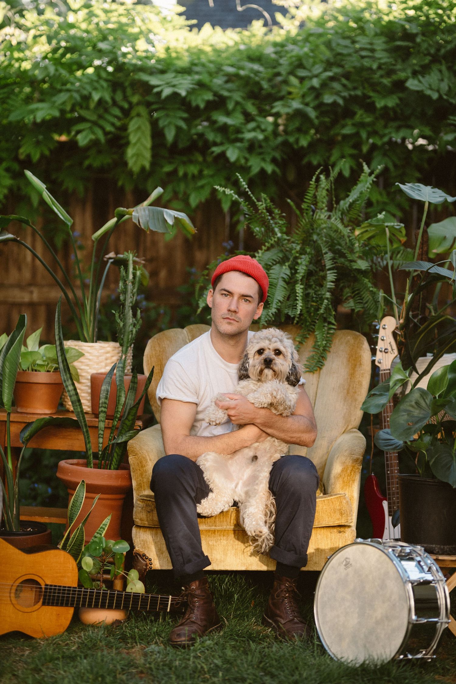 Musician with small dog sitting in garden chair surrounded by plants and instruments.