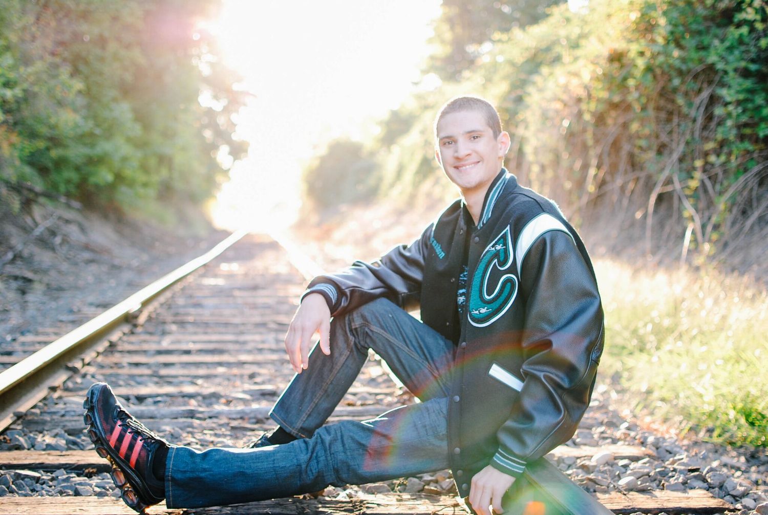 Senior photo of student in letterman jacket sitting on railroad tracks at sunset.