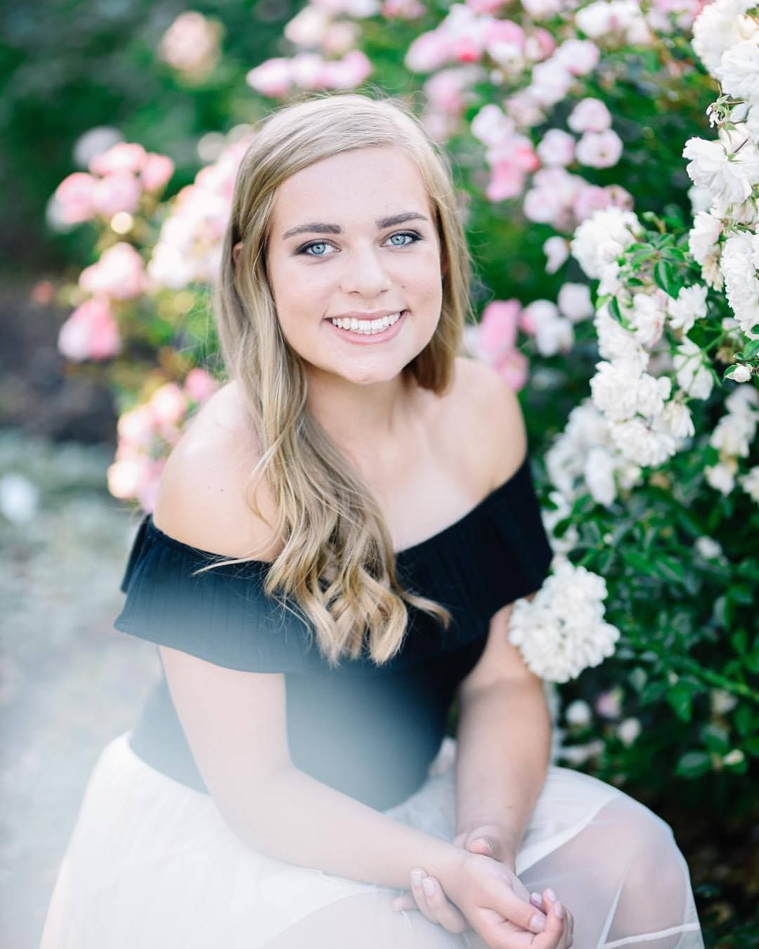 Woman in black off-shoulder top poses next to blooming pink and white roses.