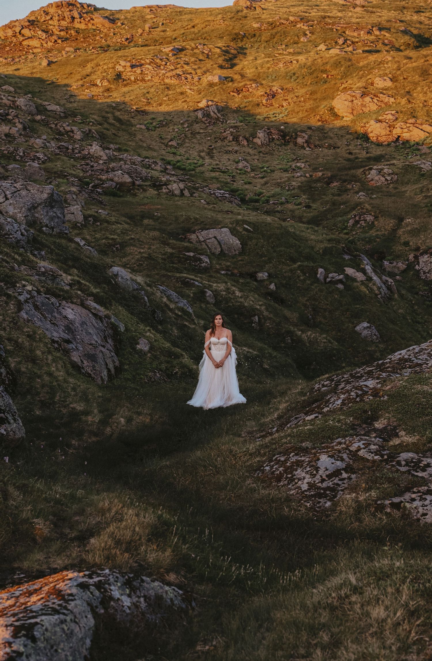 A person in a white wedding dress stands alone in a dramatic mountain landscape at golden hour.
