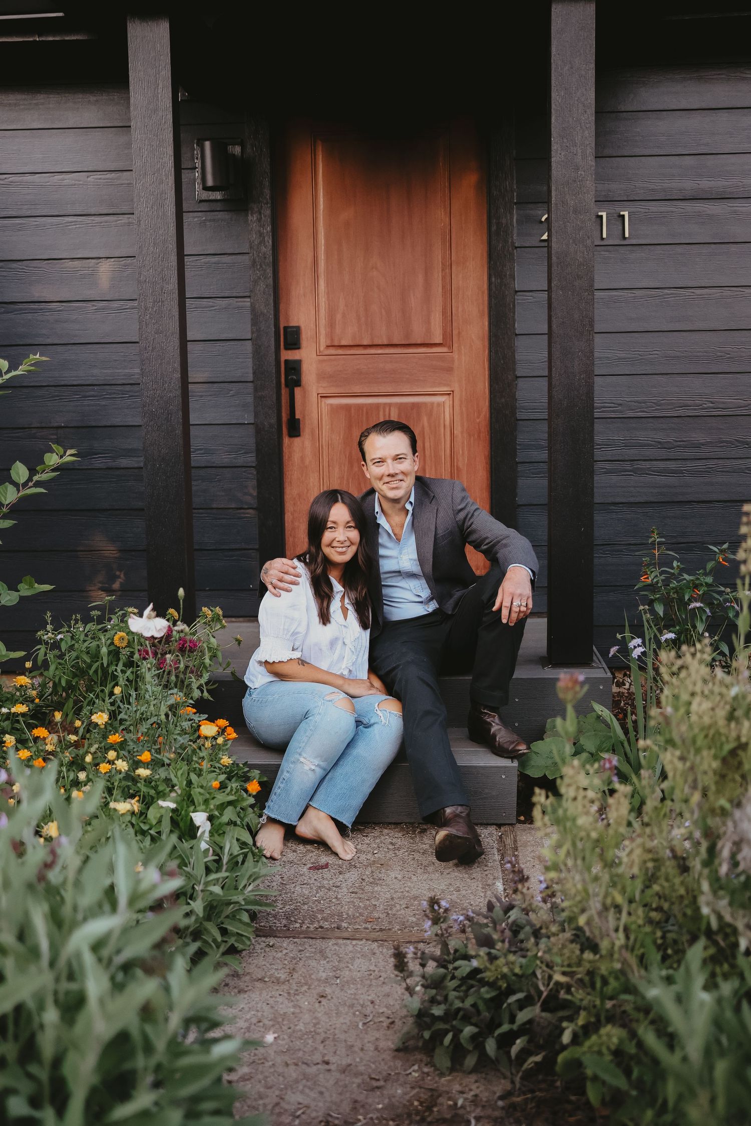 A couple sits together on steps in front of a rustic brown door surrounded by garden flowers.