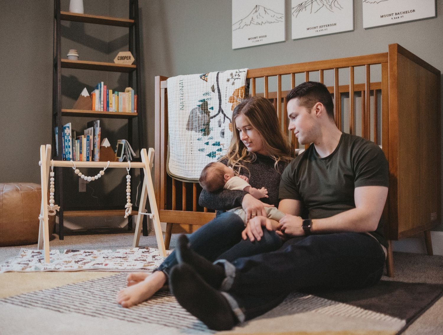 Parents sitting on floor with infant in modern nursery with artwork and wooden crib.