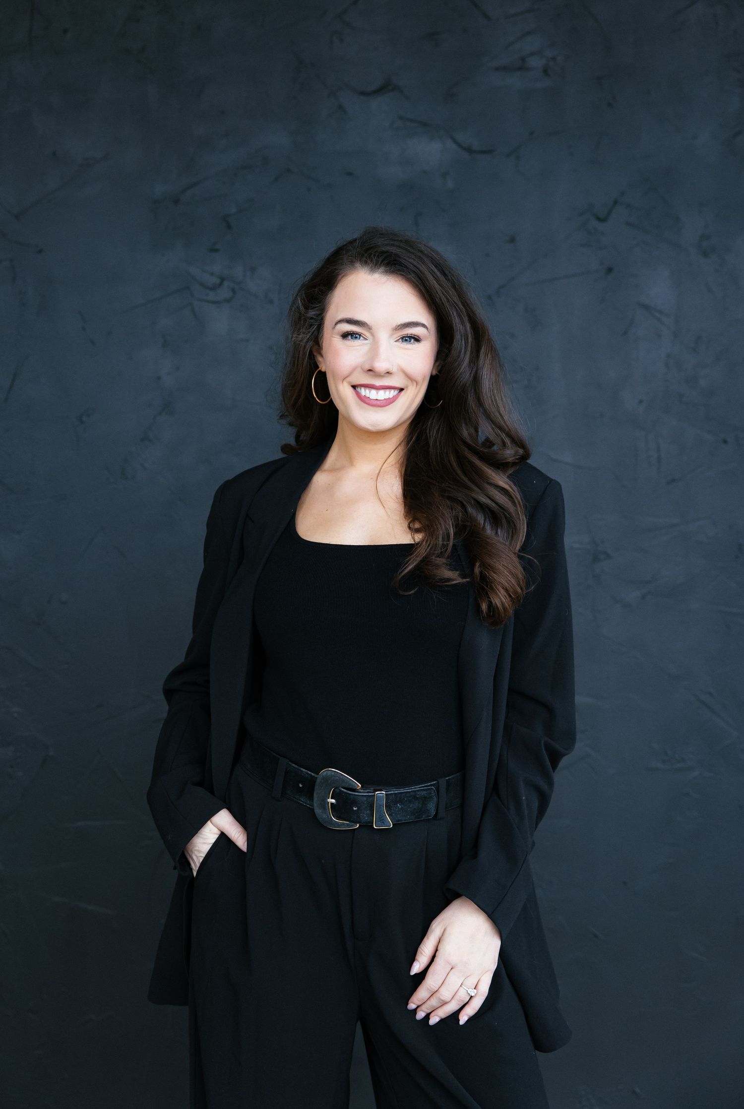 Smiling woman in all-black outfit with blazer and belt poses confidently against dark textured background.