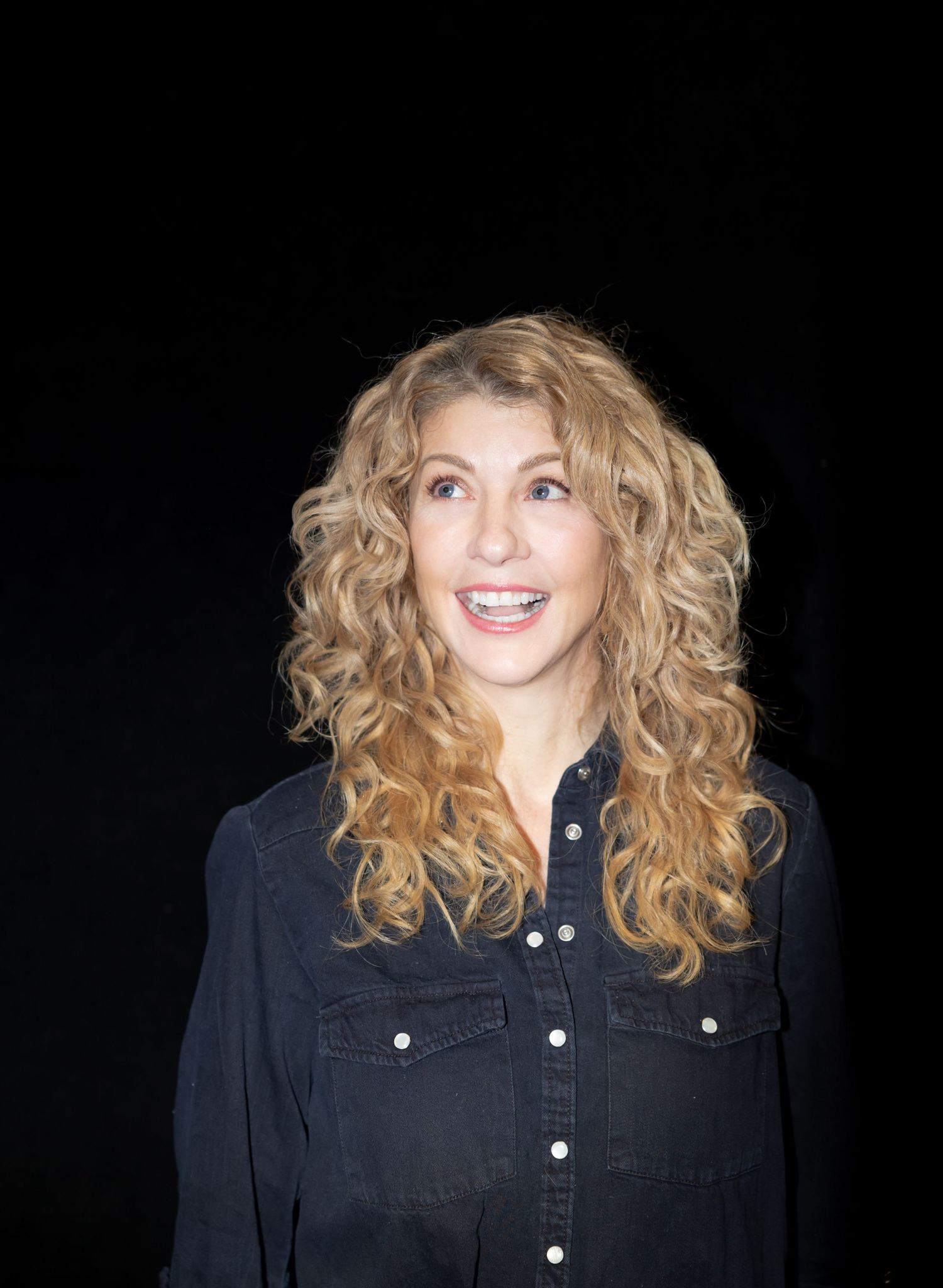 Professional headshot showing curly blonde hair and denim shirt against dark backdrop.