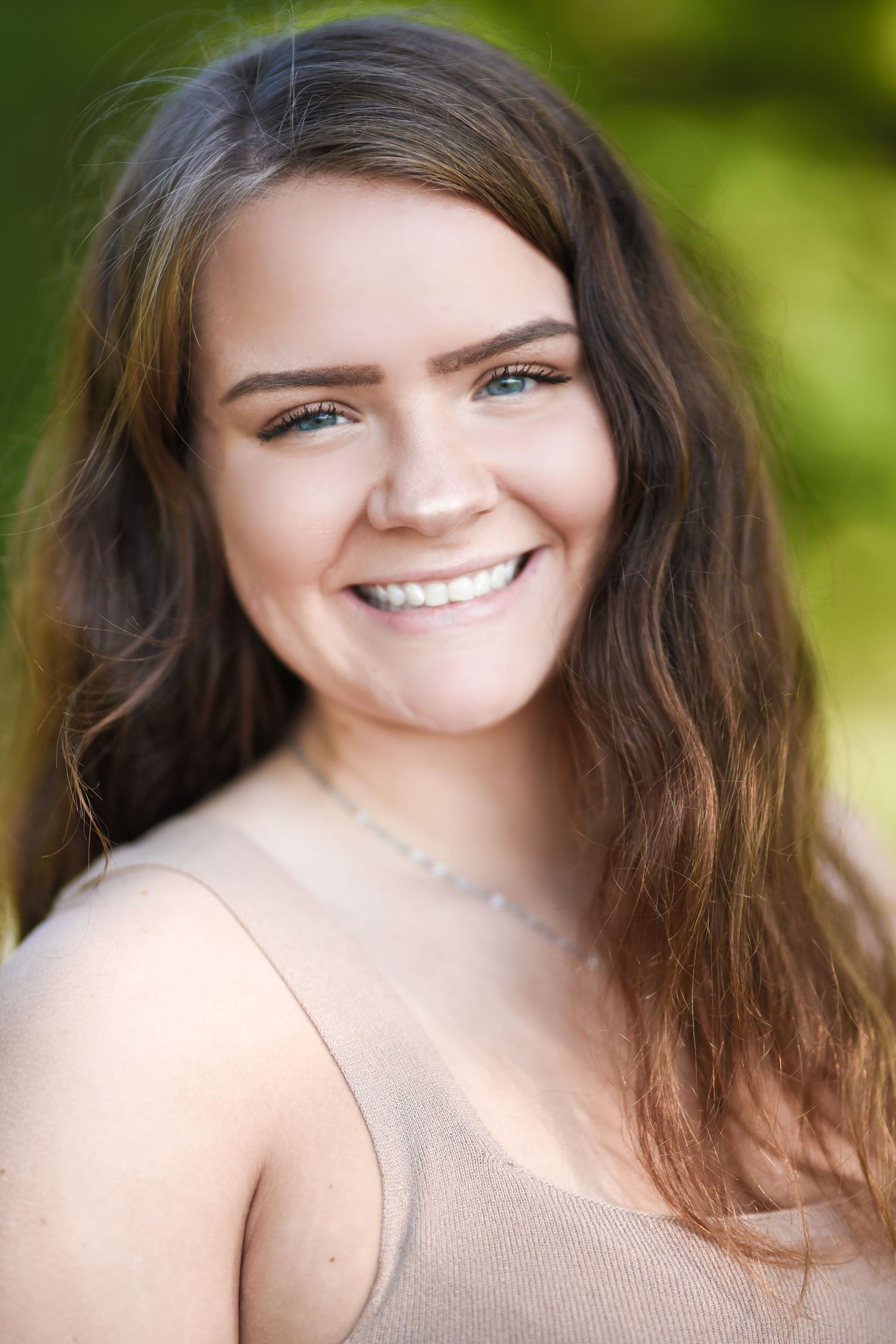 Portrait of smiling woman with long brown hair wearing beige tank top against blurred green background.