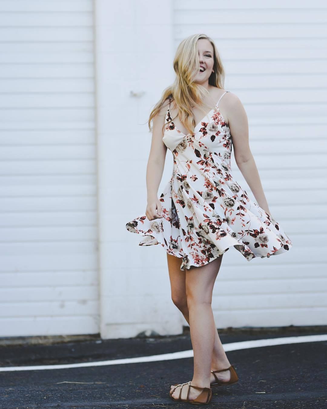 Woman in floral sundress twirling playfully against white wall background.