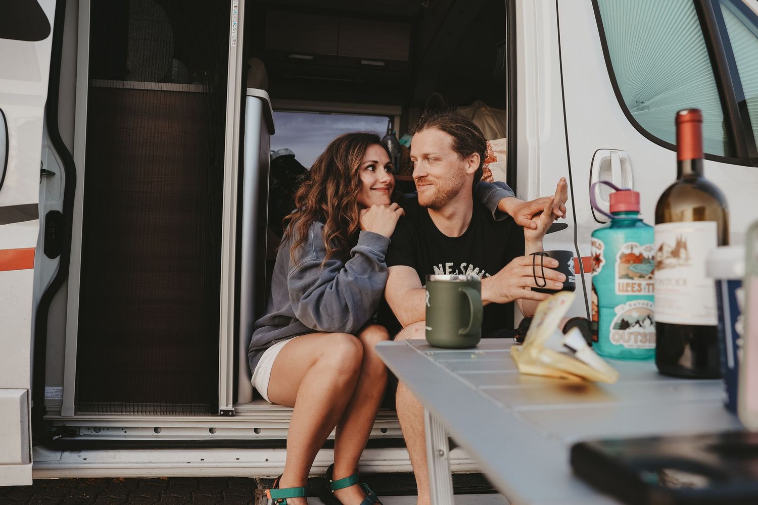 A couple enjoys drinks and snacks while relaxing in their camper van.