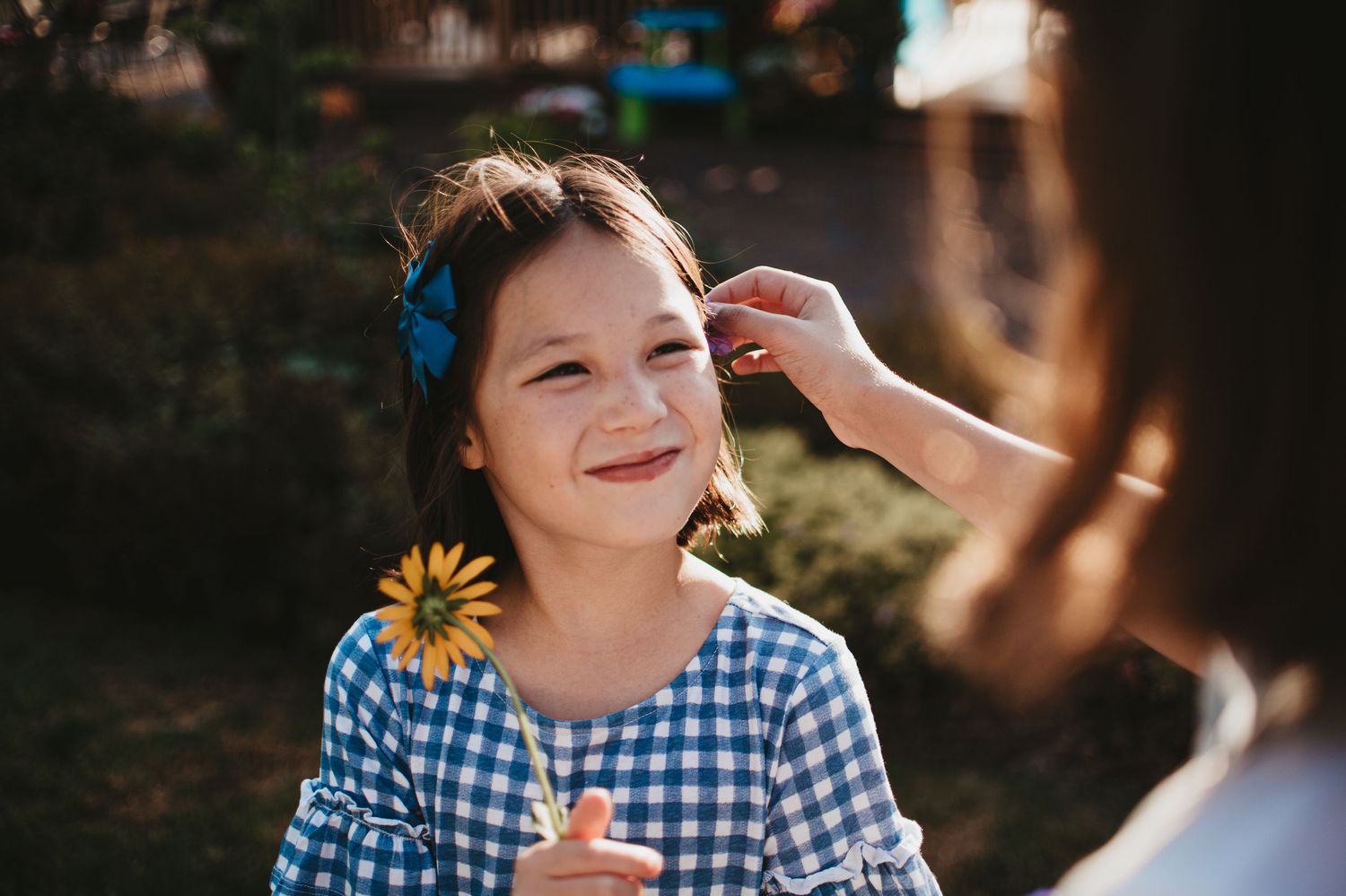 Child in blue gingham dress holds sunflower while enjoying a sunny day outdoors.