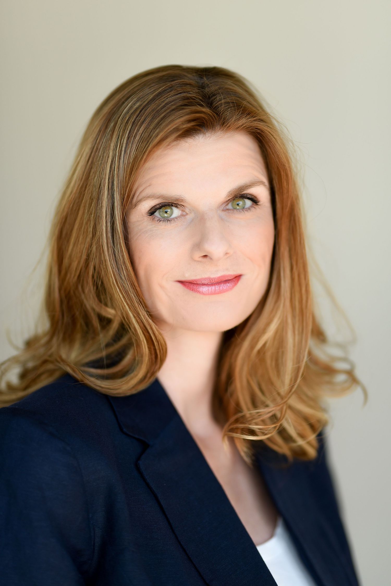 Professional headshot of a person in a navy blazer with styled hair against a light background.