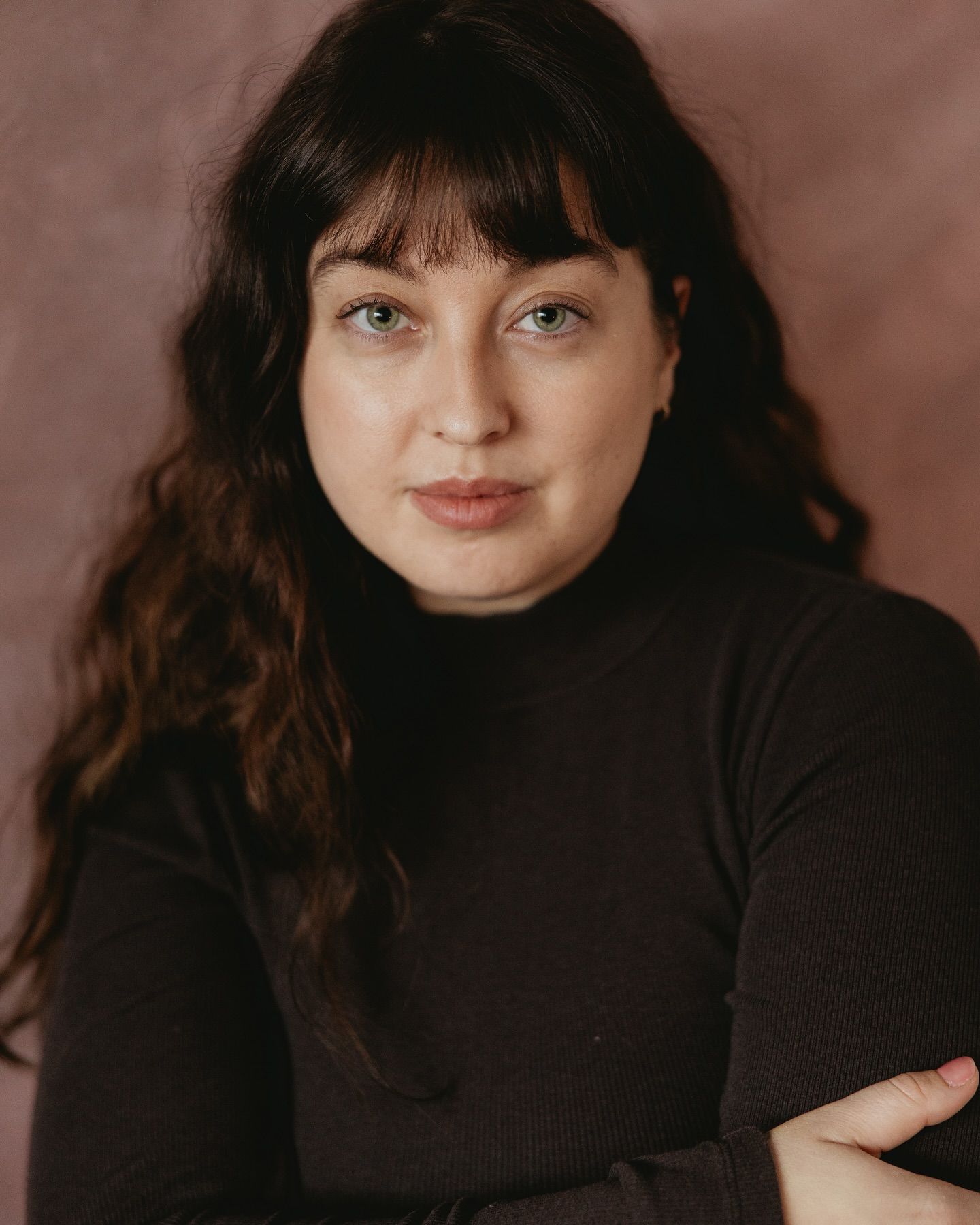 Professional portrait of person in black turtleneck sweater with long dark wavy hair against brown background.