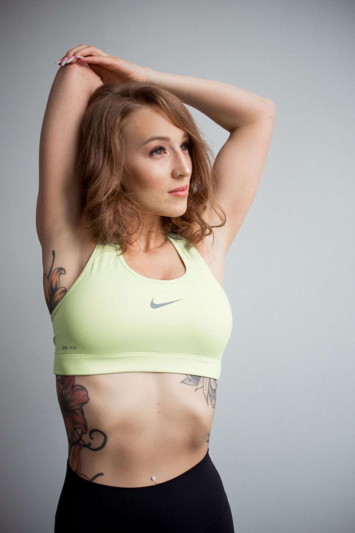 Athletic figure in yellow sports bra with visible tattoos posing against grey background.