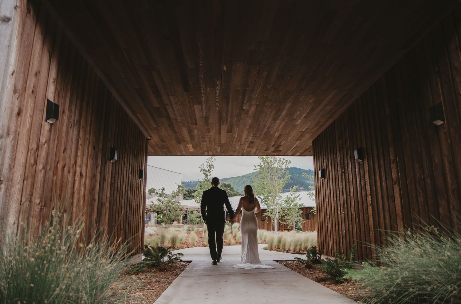 A couple walking through a wooden covered walkway surrounded by greenery.