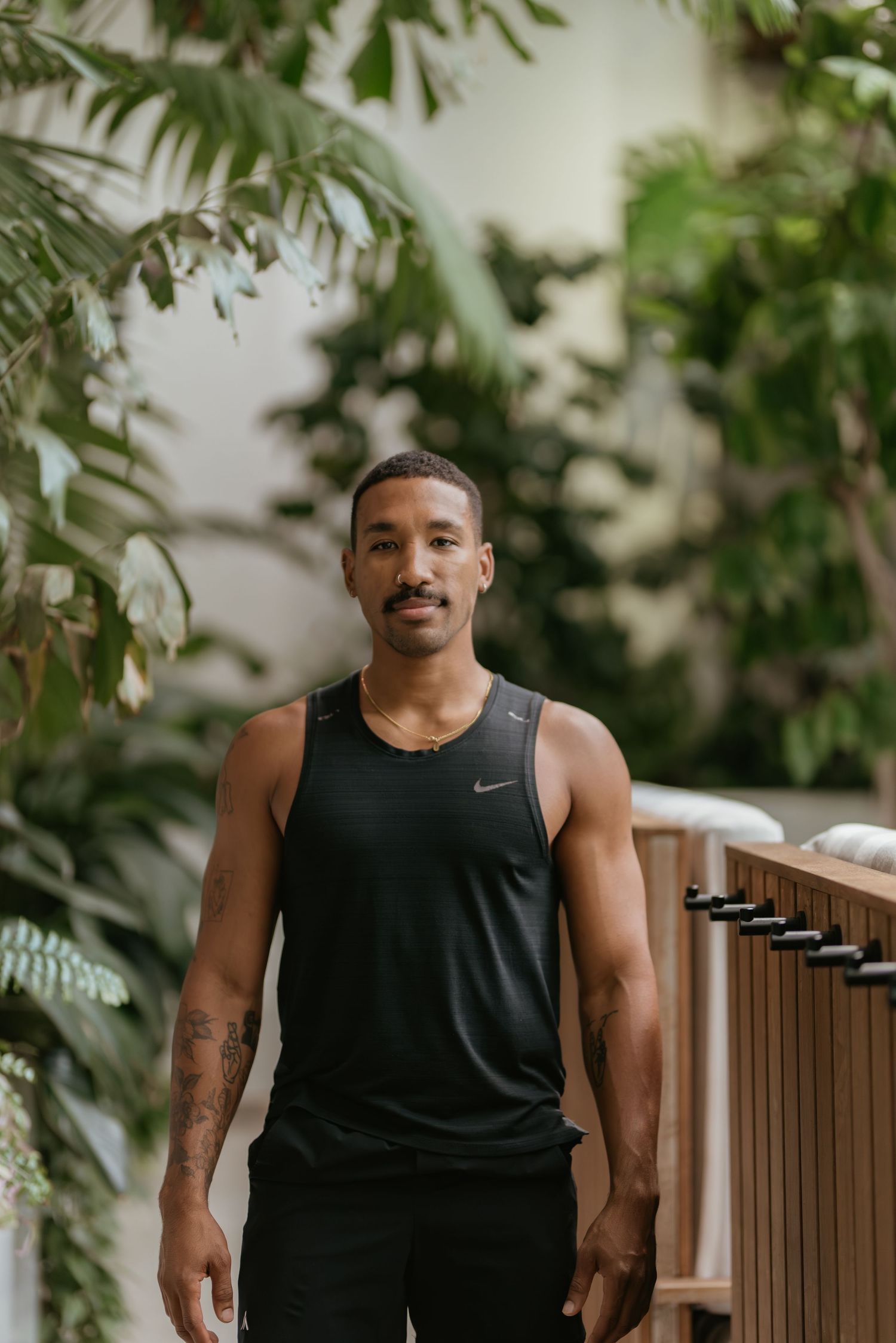 Athletic man in black Nike tank top standing among lush green tropical plants in a modern indoor setting.