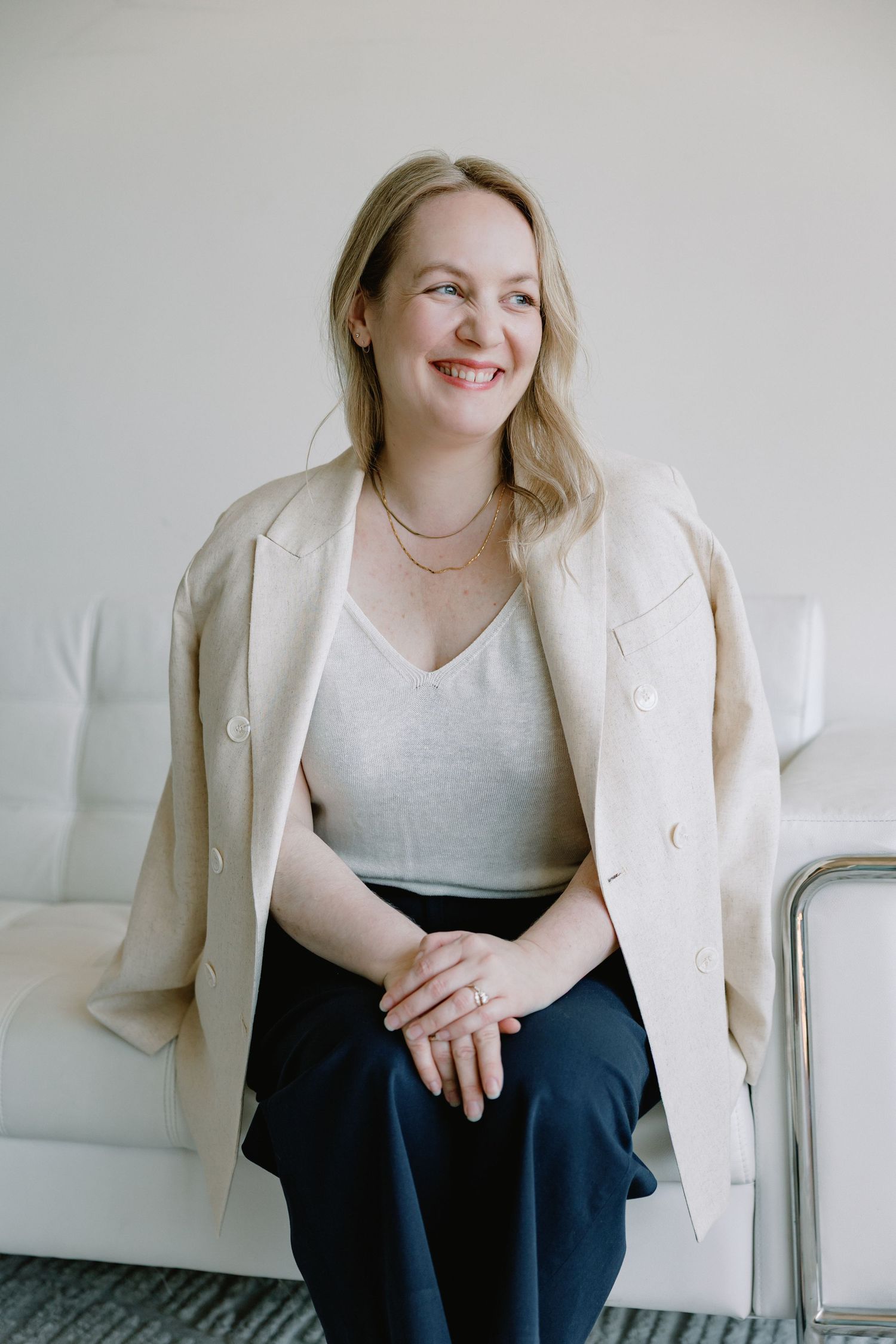 Smiling blonde woman in beige blazer and gray top sits on white sofa in bright minimalist setting.