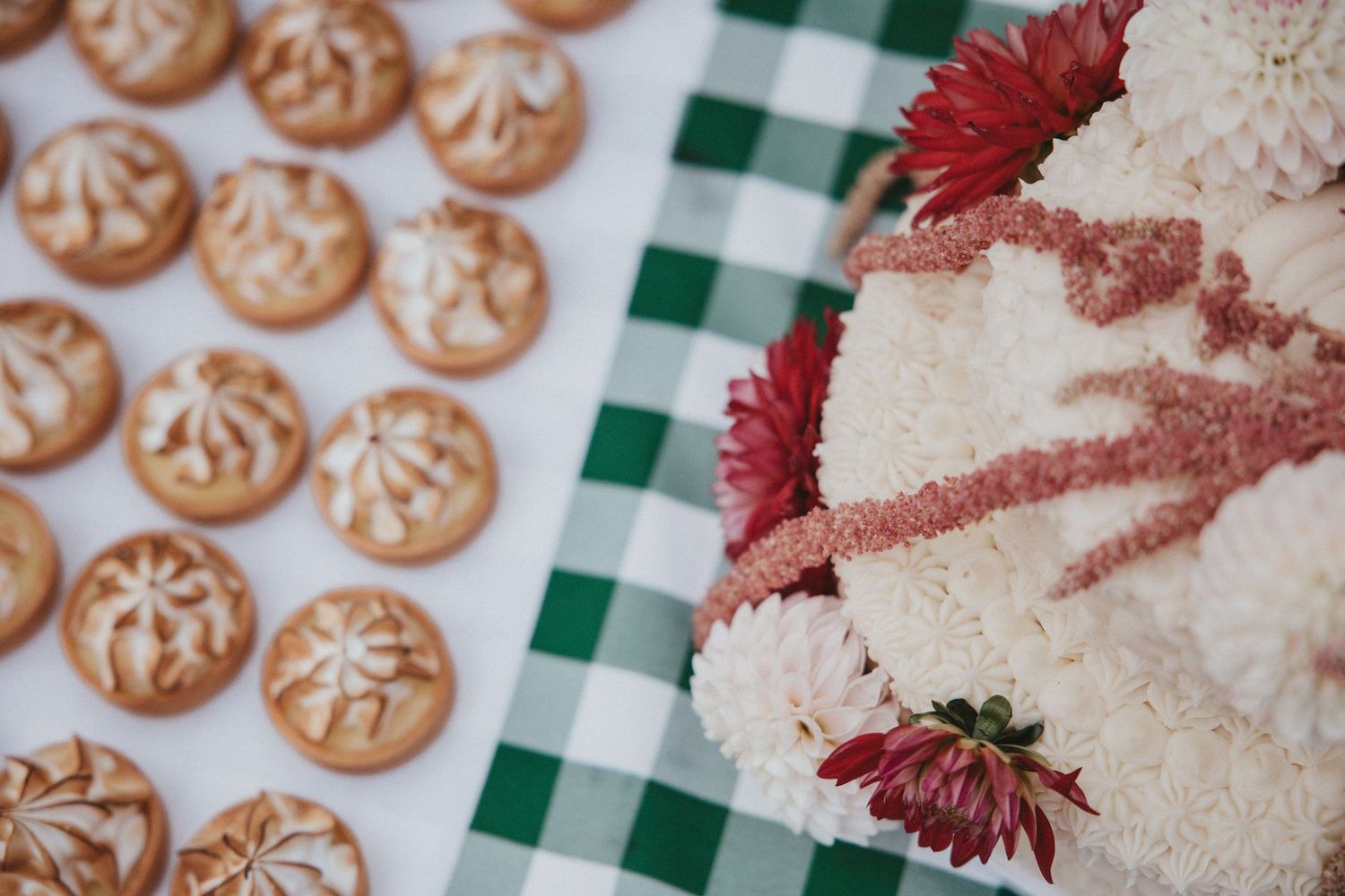 Rows of decorated Christmas sugar cookies and red flowers on a green checkered tablecloth.