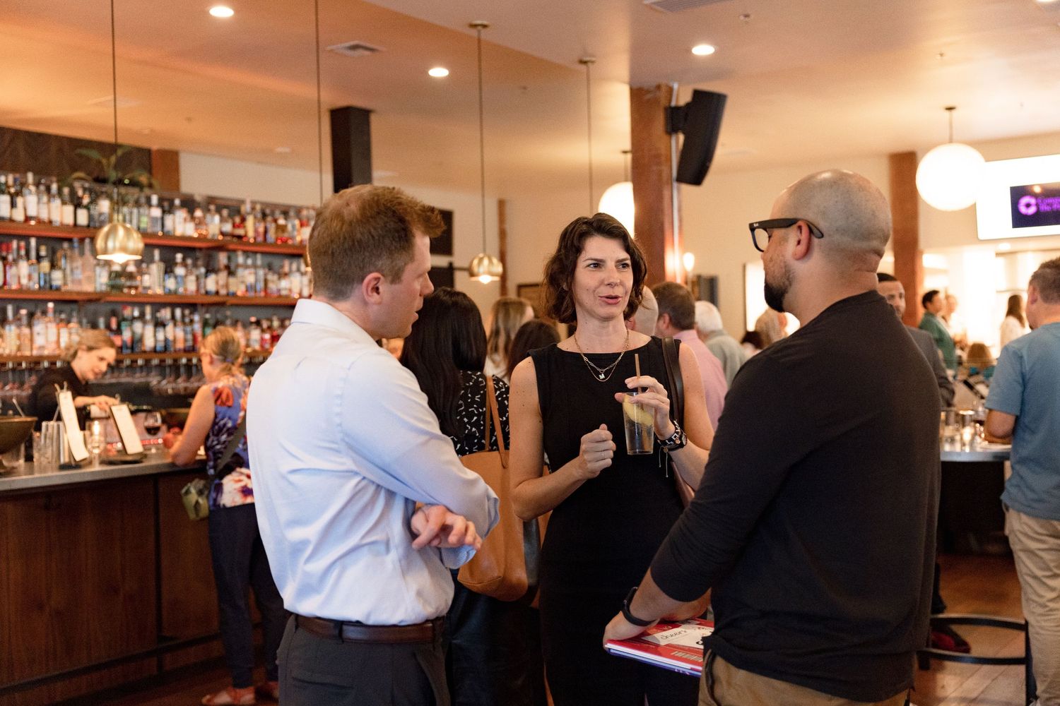 Patrons sit at a long bar counter inside a dimly lit restaurant with bottles displayed on shelves behind the bar.